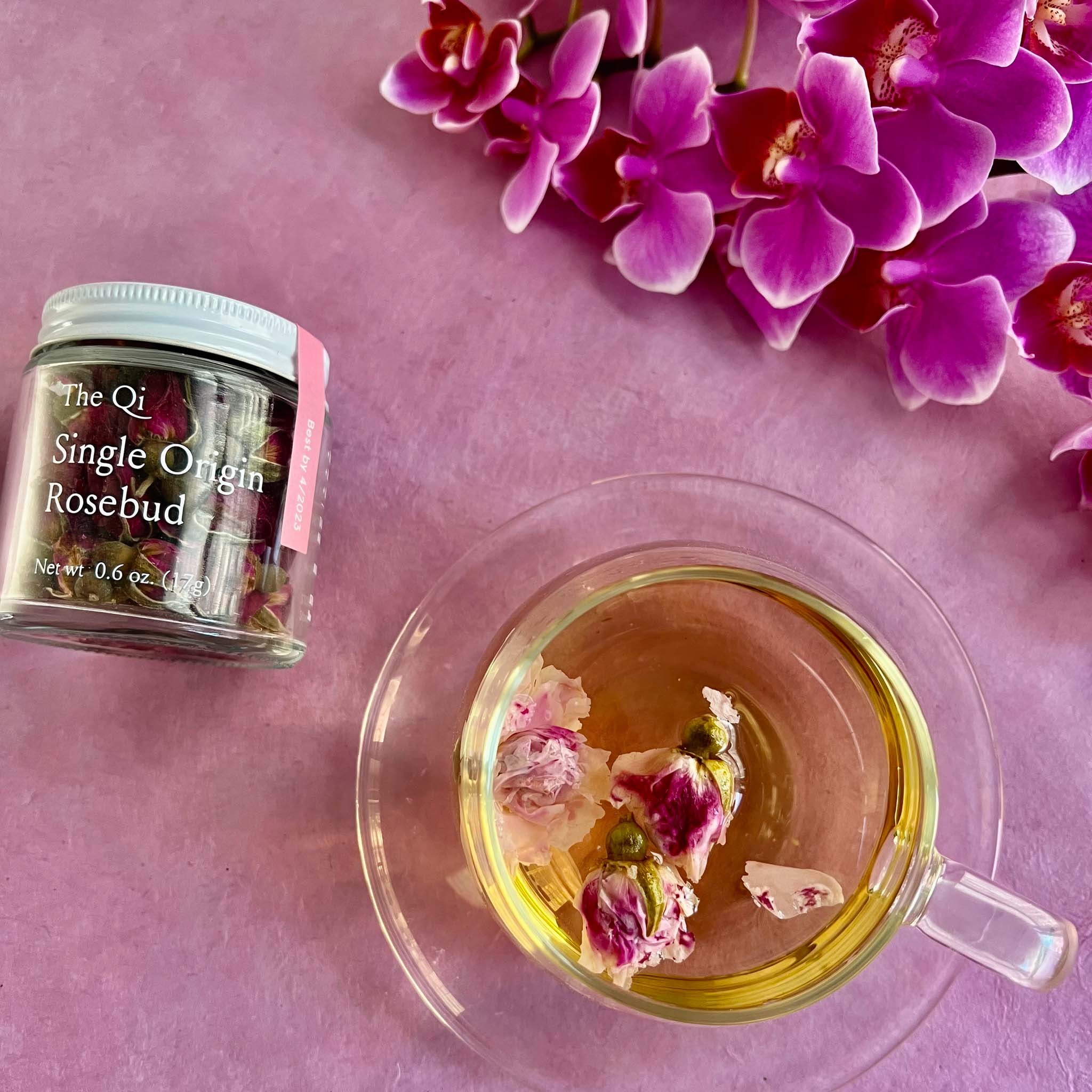 Tea cup with rosebud tea, jar labeled 'The Qi Single Origin Rosebud', and pink flowers on a pink background