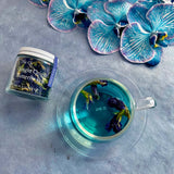 Tea cup with butterfly pea tea, jar labeled 'The Qi Single Origin Butterfly Pea', and blue and white flowers on a blue background