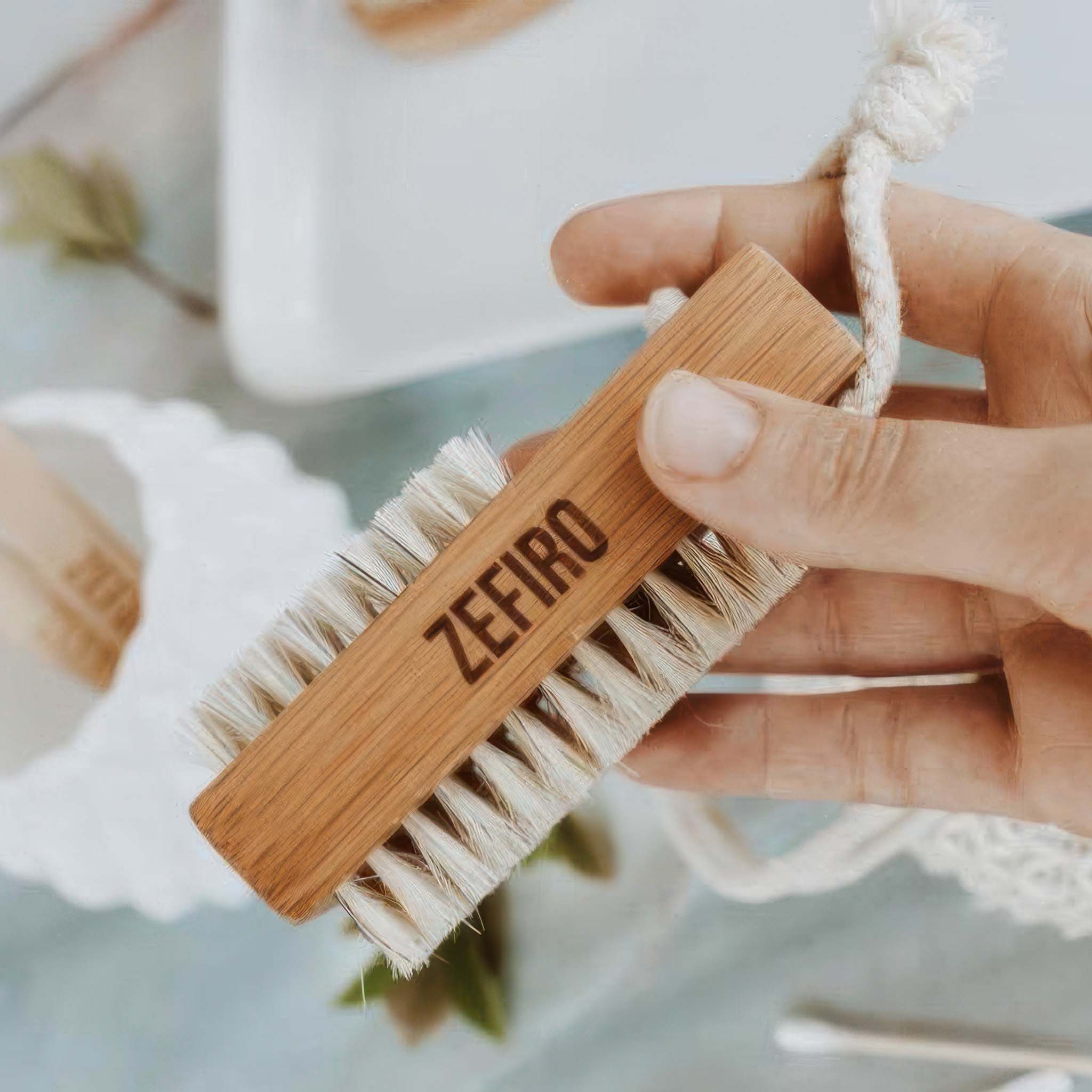 Hand holding a wooden nail brush with bristles labeled 'Zefiro' against a blurred background.