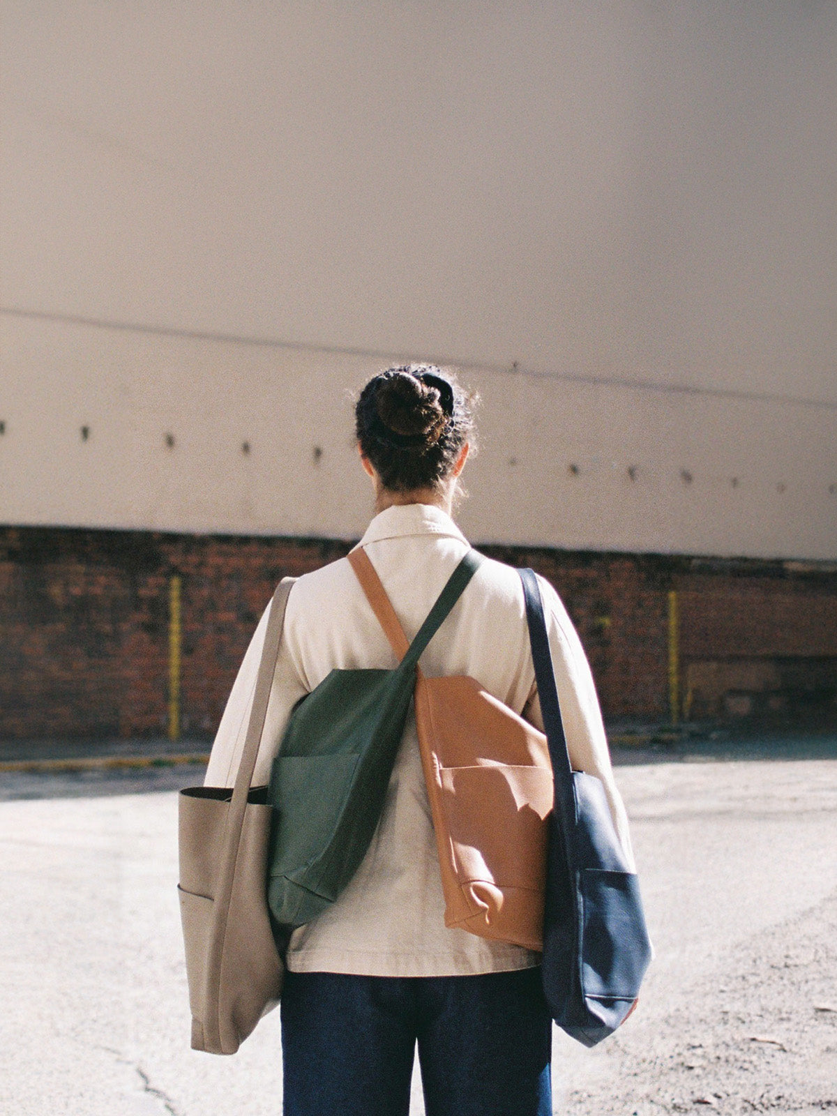 Model standing outdoors with their back to the camera, wearing a light jacket and carrying multiple tote bags in beige, green, tan, and navy.