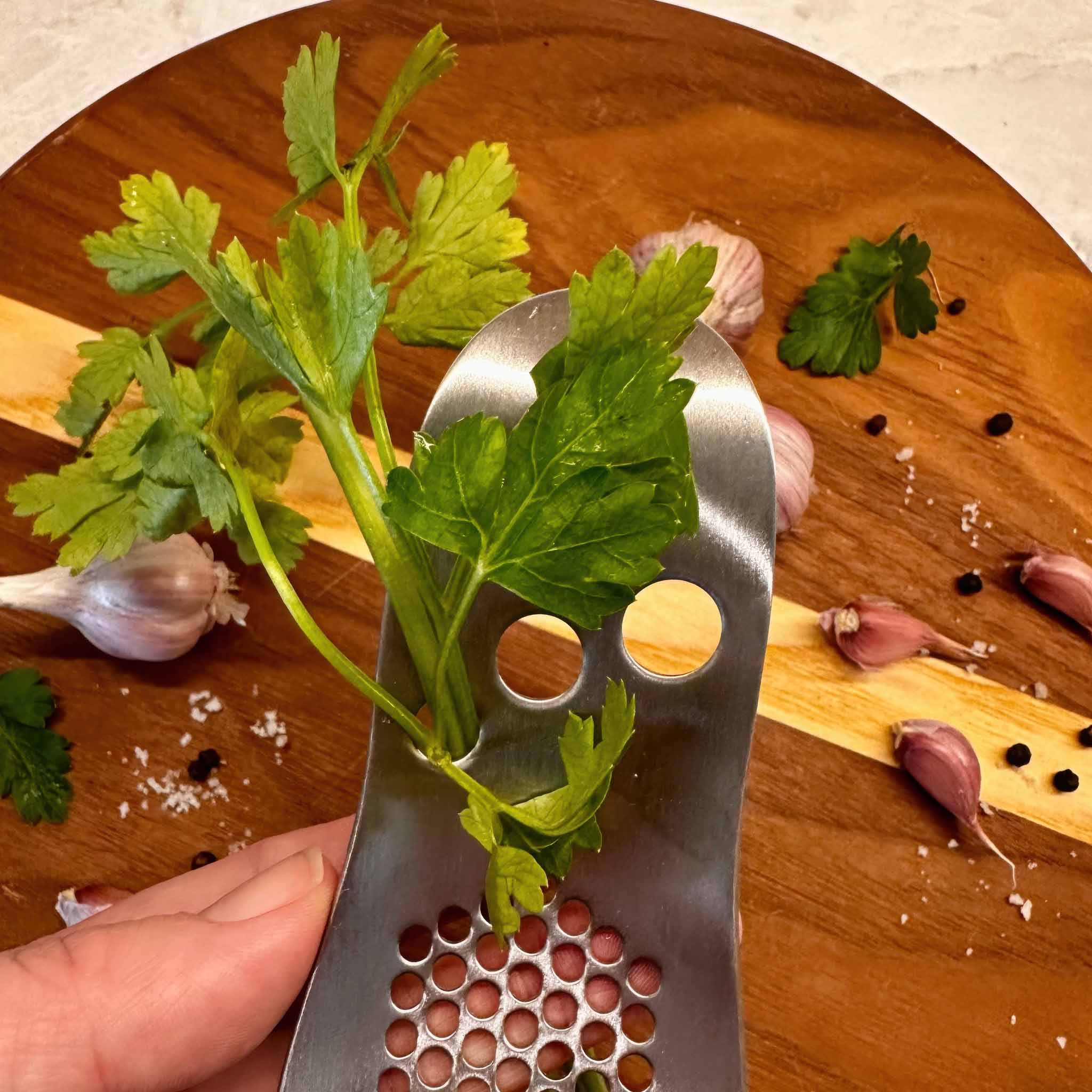Metallic herb stripper with parsley on a wooden board with garlic and herbs.