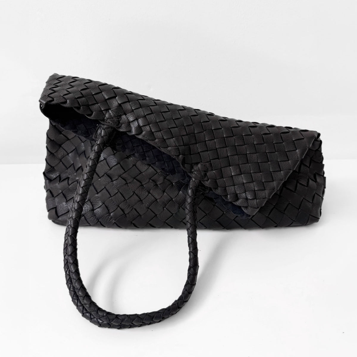 A black woven leather bag toppled over with a matching braided shoulder strap, photographed on a clean white studio background.