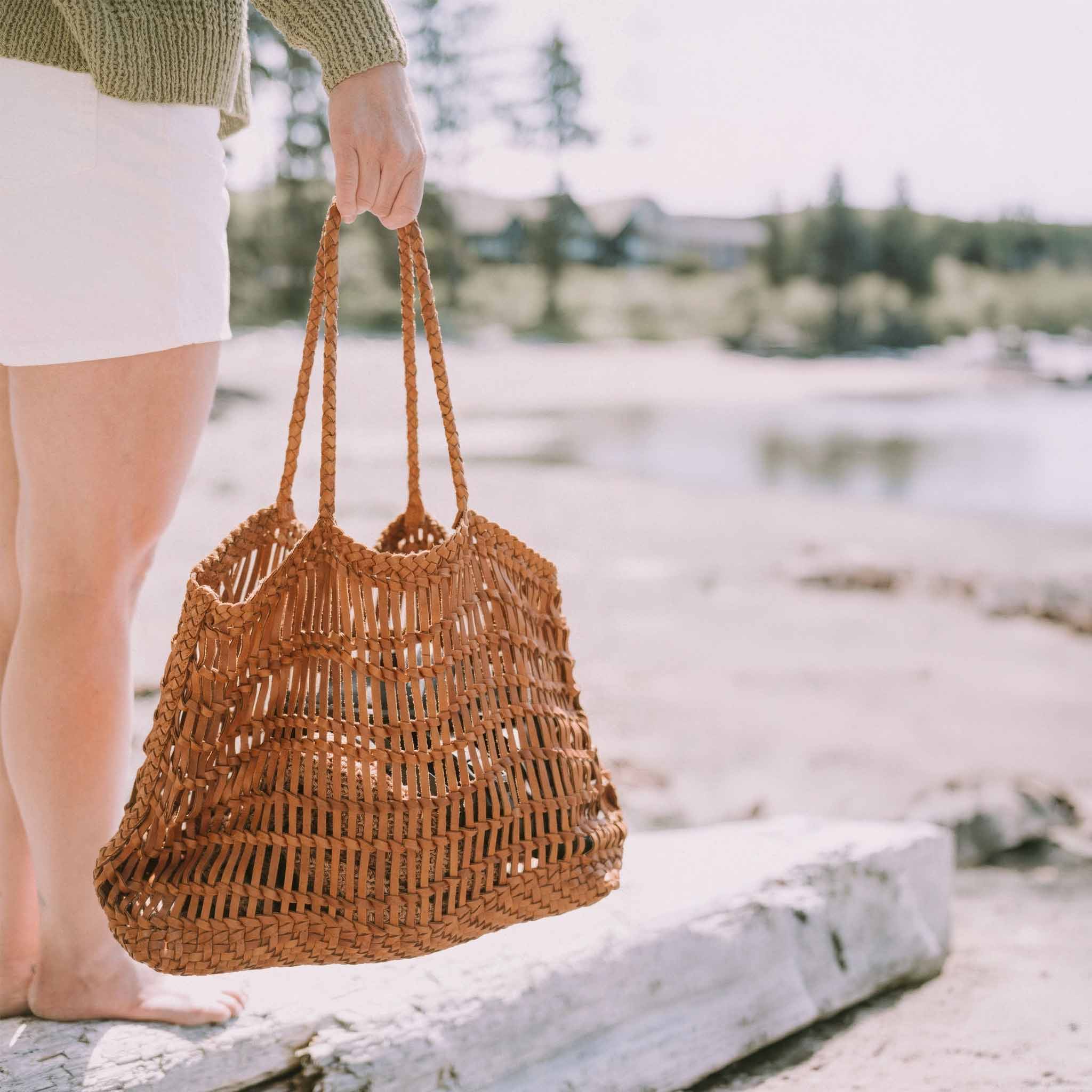 Person holding a woven brown bag by a body of water with trees in the background