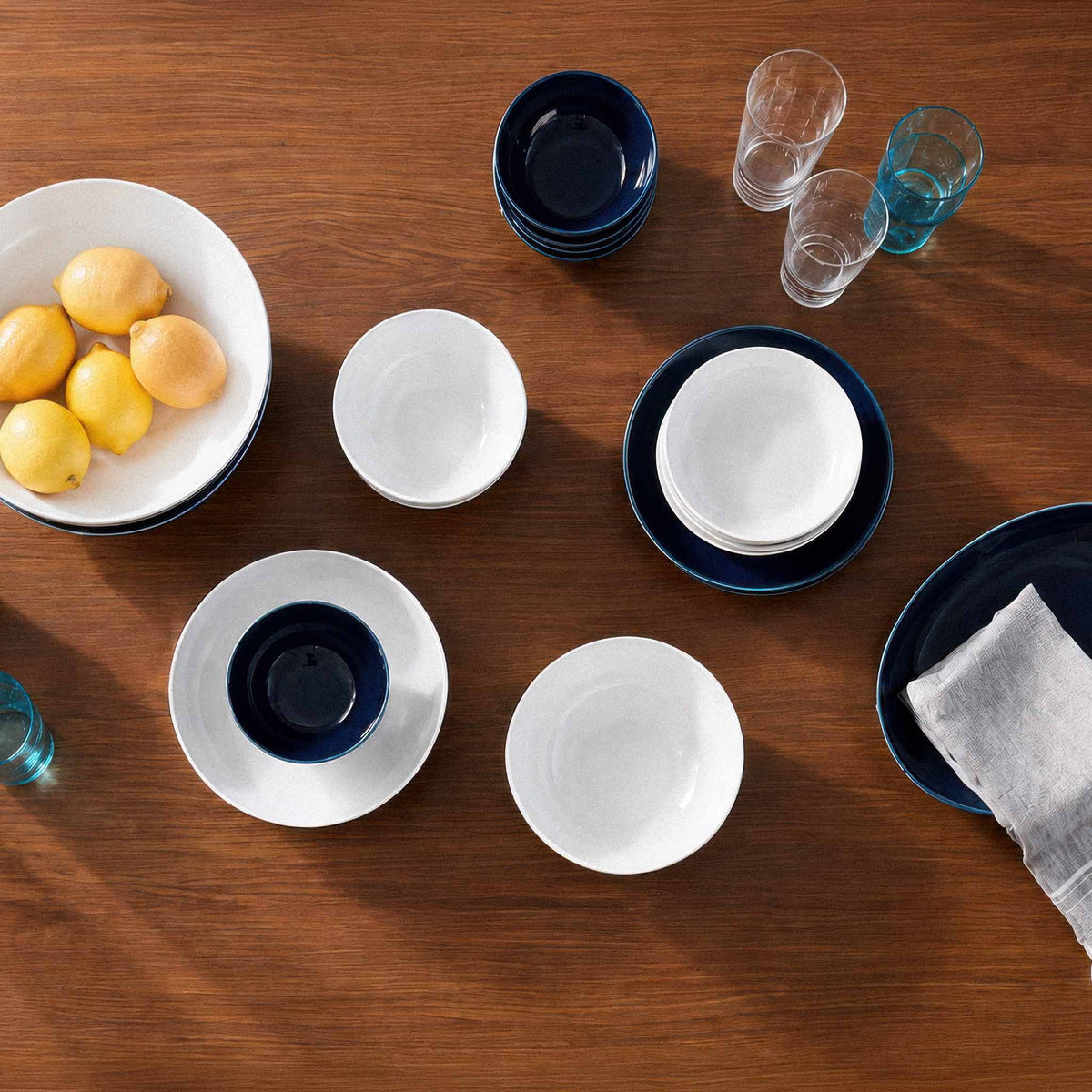 A flat-lay view of a dinnerware set on a wooden table, featuring white ceramic bowls, plates, and deep navy blue plates and bowls, styled with lemons, drinking glasses, and a napkin.