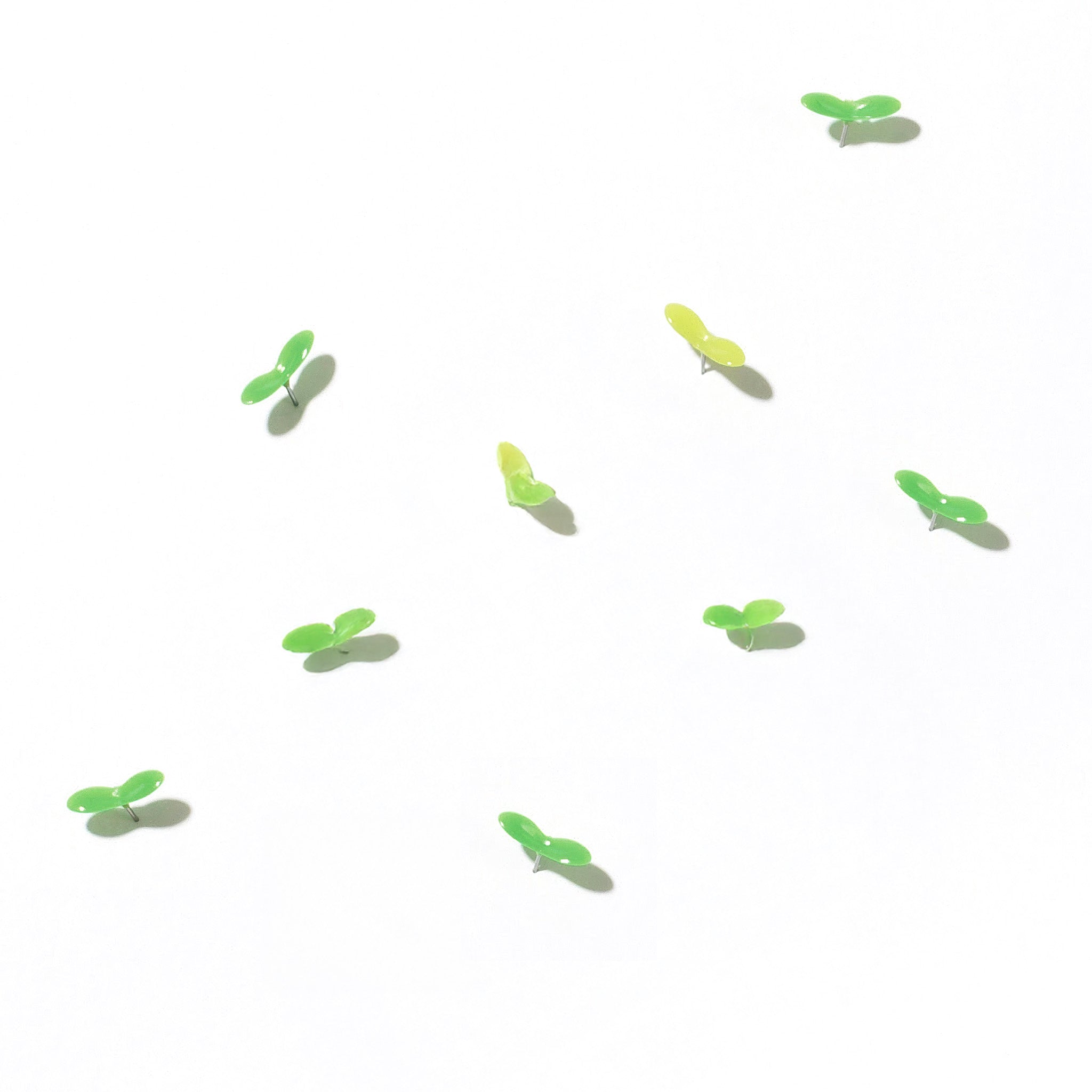 A scattering of small green push pins shaped like sprouting leaves arranged on a white surface, creating a minimalist and playful design reminiscent of new plant growth.