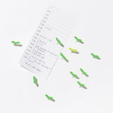 A minimalist desk scene with bright green leaf-shaped push pins securing a handwritten daily schedule on a white surface. The playful pins resemble small sprouts, adding a natural, lively touch to the clean workspace layout.
