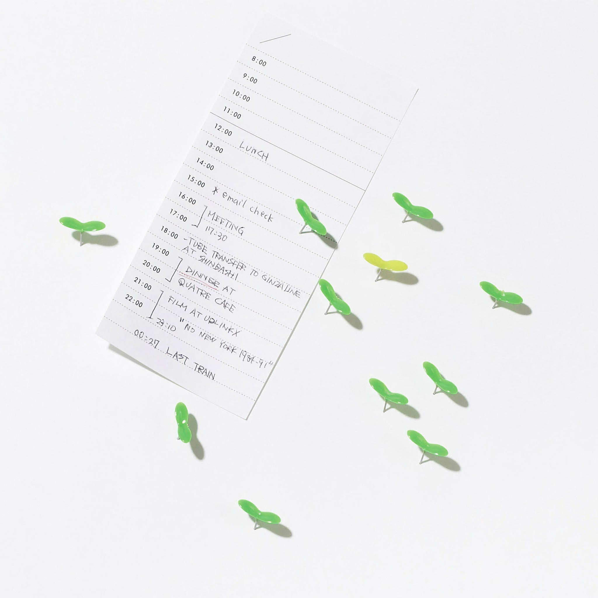 A minimalist desk scene with bright green leaf-shaped push pins securing a handwritten daily schedule on a white surface. The playful pins resemble small sprouts, adding a natural, lively touch to the clean workspace layout.