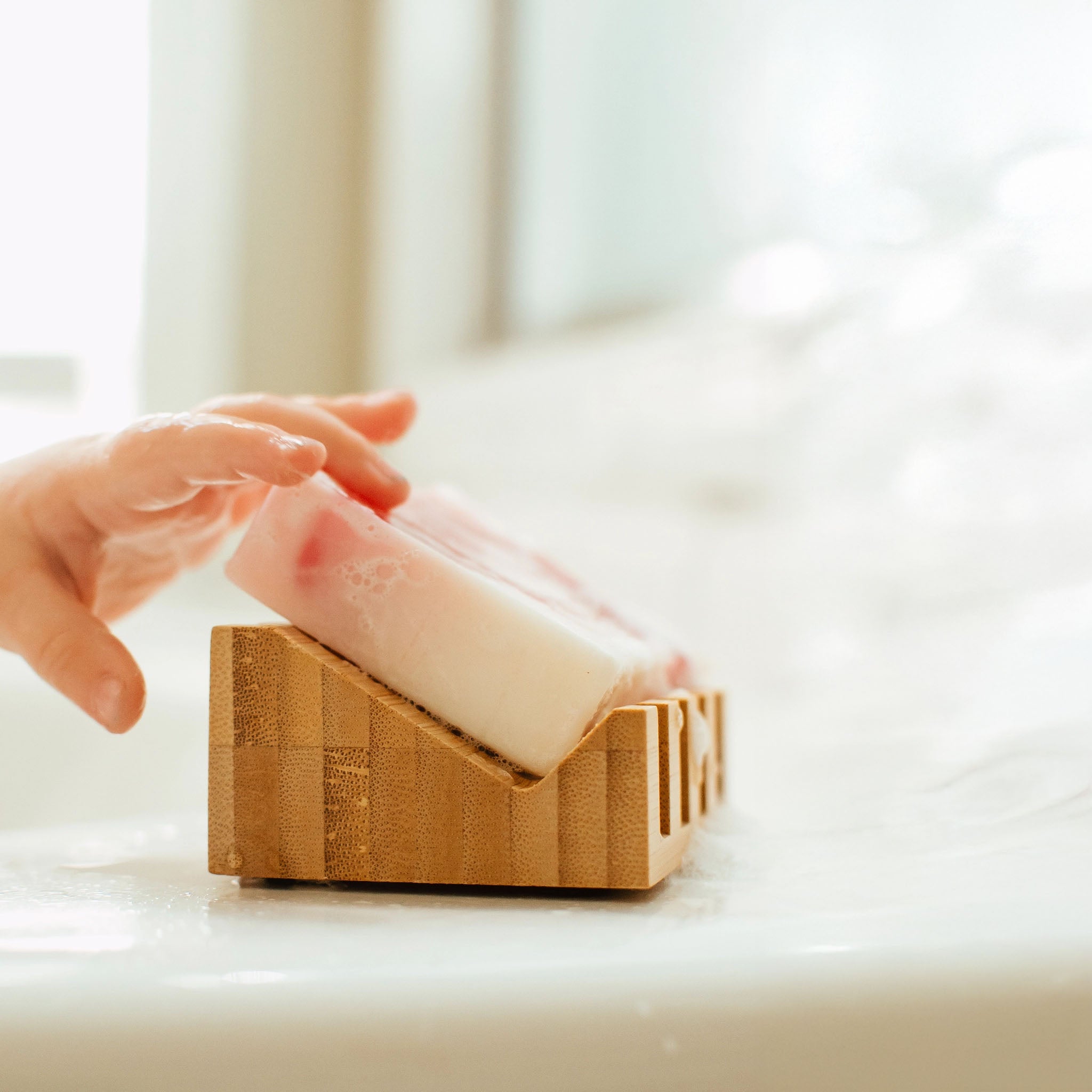 Hand reaching for a bar of soap in a wooden holder on a light surface.
