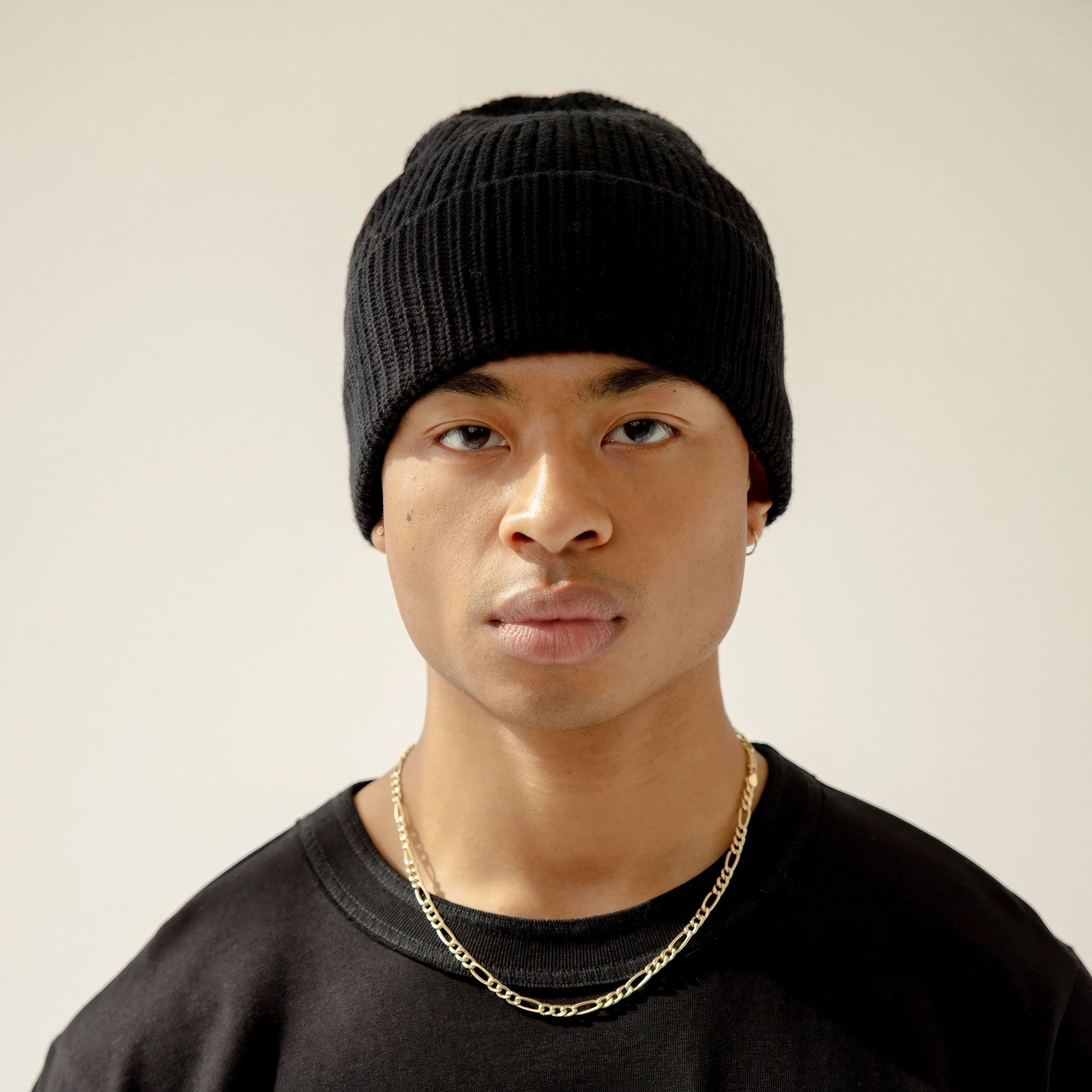 Model wearing a black knit beanie and black shirt with a gold chain on a plain background