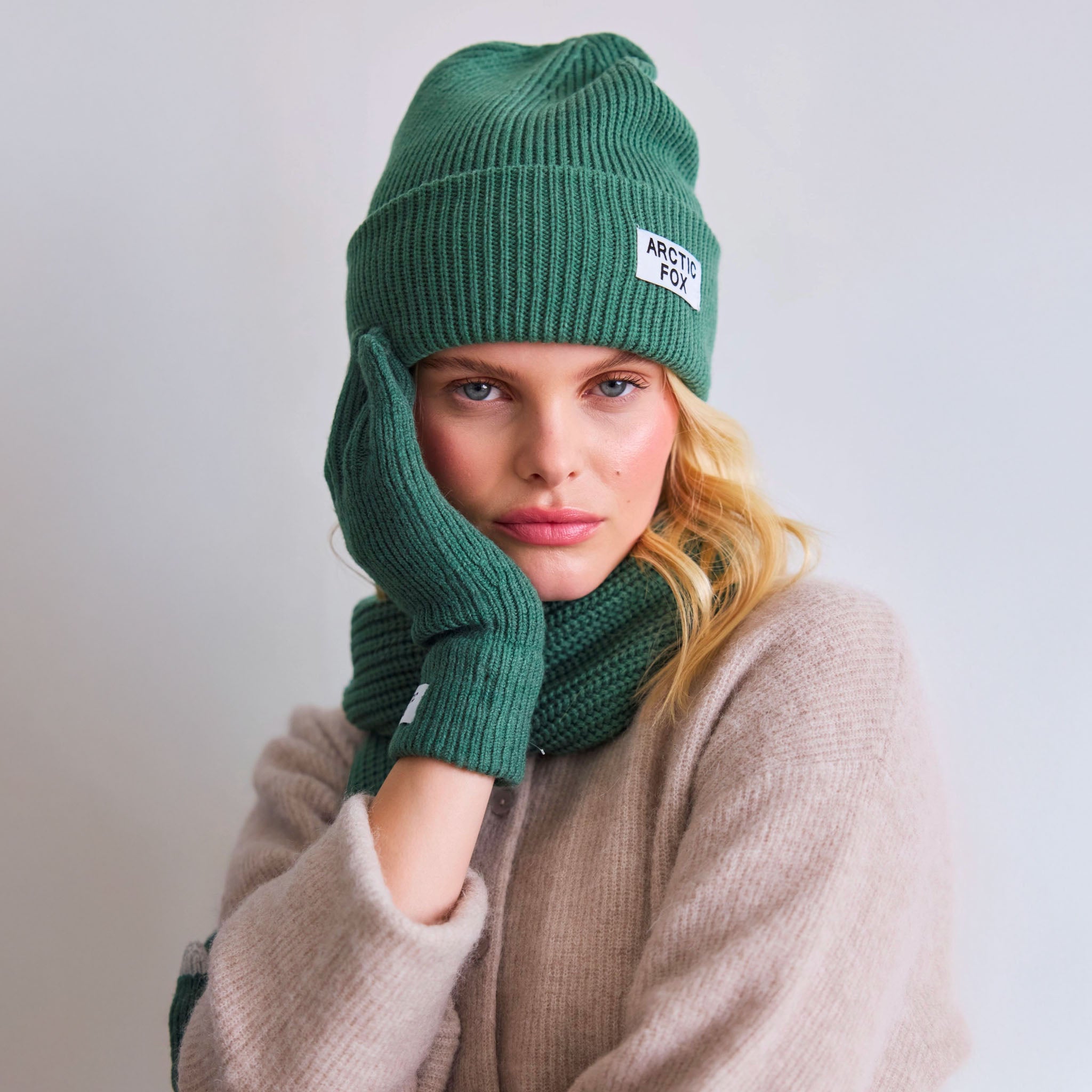 Person wearing a green knit hat and scarf with 'Arctic Fox' branding on a light gray background