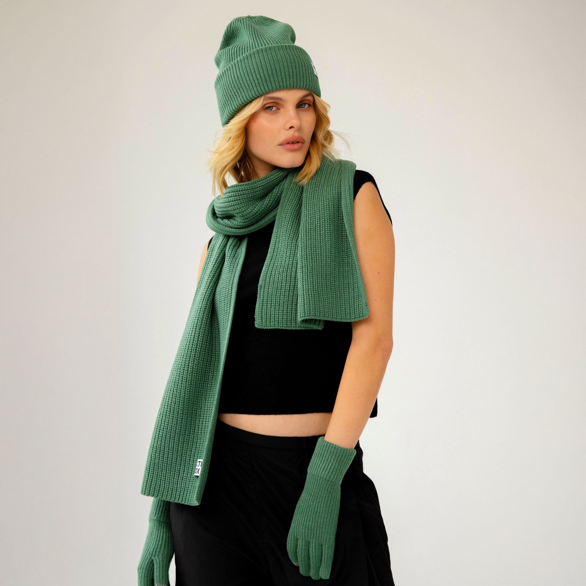 Woman wearing a fern green knit hat, scarf, and gloves on a plain background