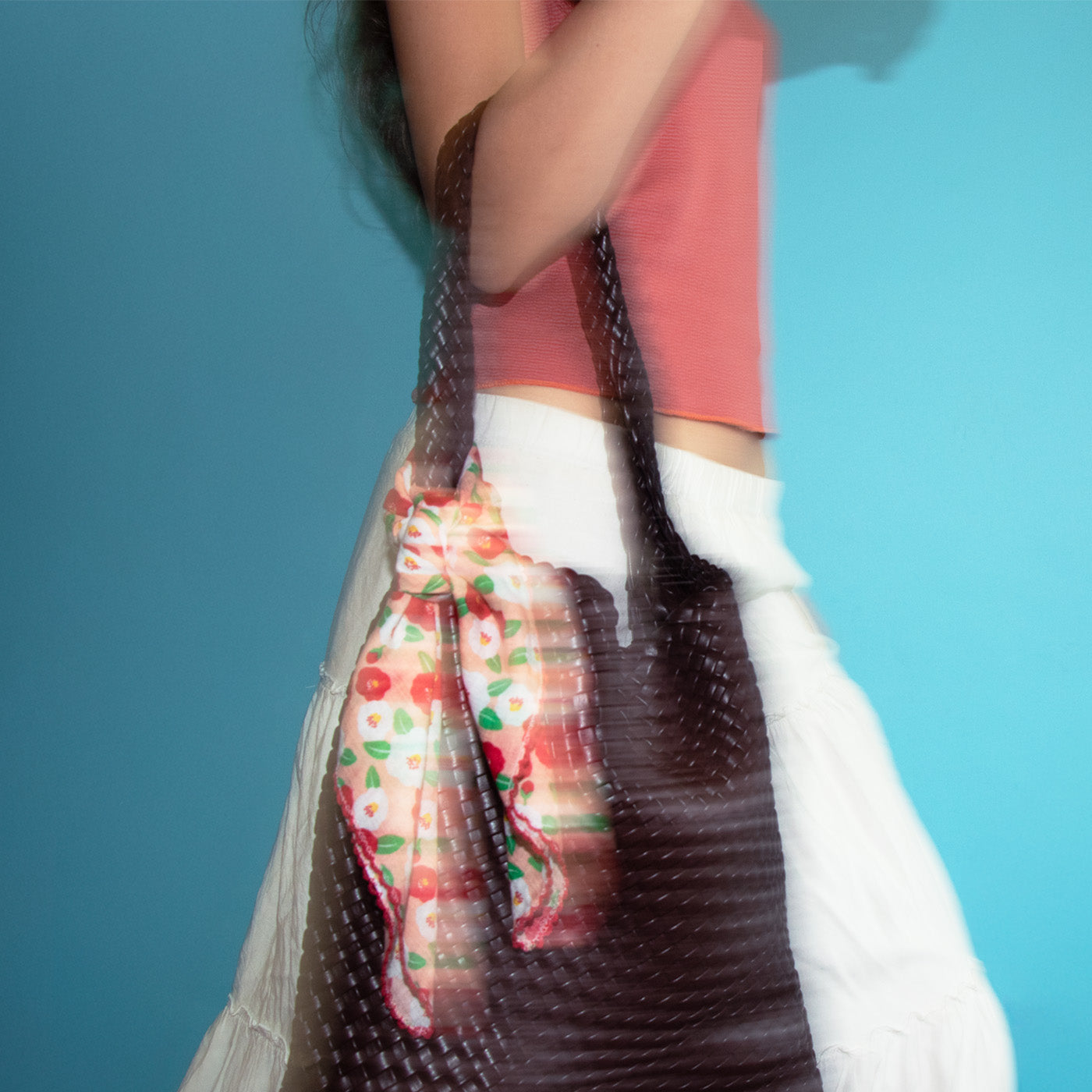 Model is carrying a dark brown bag with a patterned bandana draped around bag against a blue background