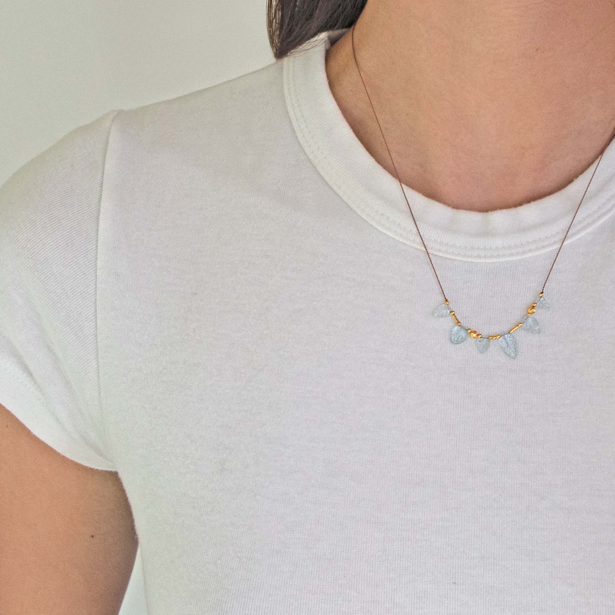 Person wearing a white T-shirt and See Real Flowers Leaf Necklace with Aquamarine + Gold beads.