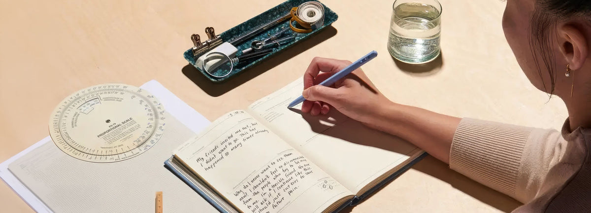 Person writing in a notebook with a pen, surrounded by stationery items on a light surface.