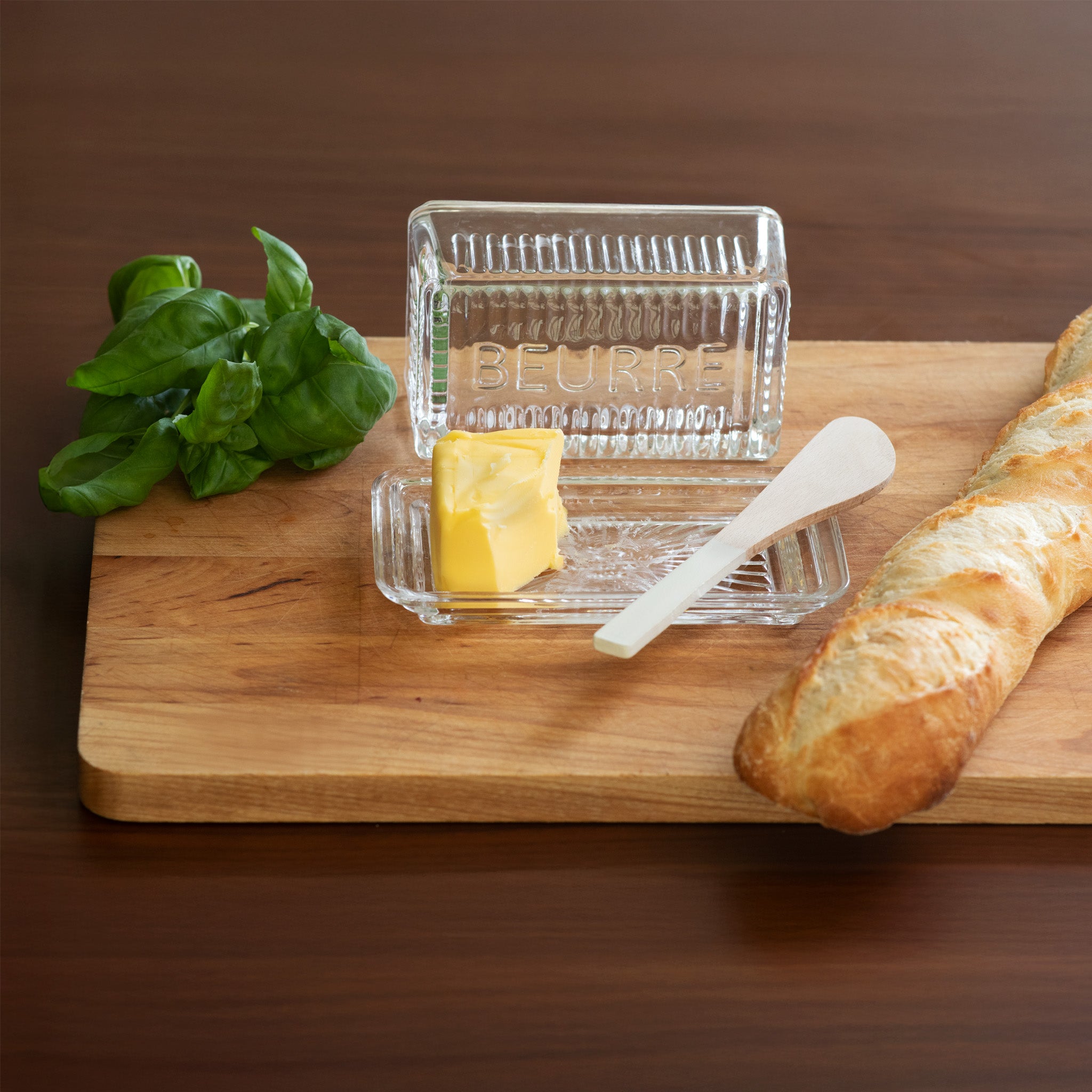 Butter dish with a stick of butter, a baguette, and basil leaves on a wooden board.