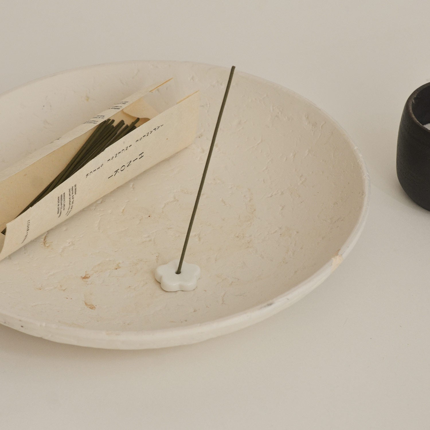 Incense stick on a white ceramic plate with a book and black container in the background.