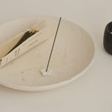 Incense stick on a white ceramic plate with a book and black container in the background.
