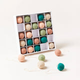 A white box containing 25 felted wool baubles in muted tones of teal, sage, cream, blush, and brown, with several ornaments scattered in front. Each round bauble has a string loop for hanging, creating a soft, natural, and handcrafted holiday look.