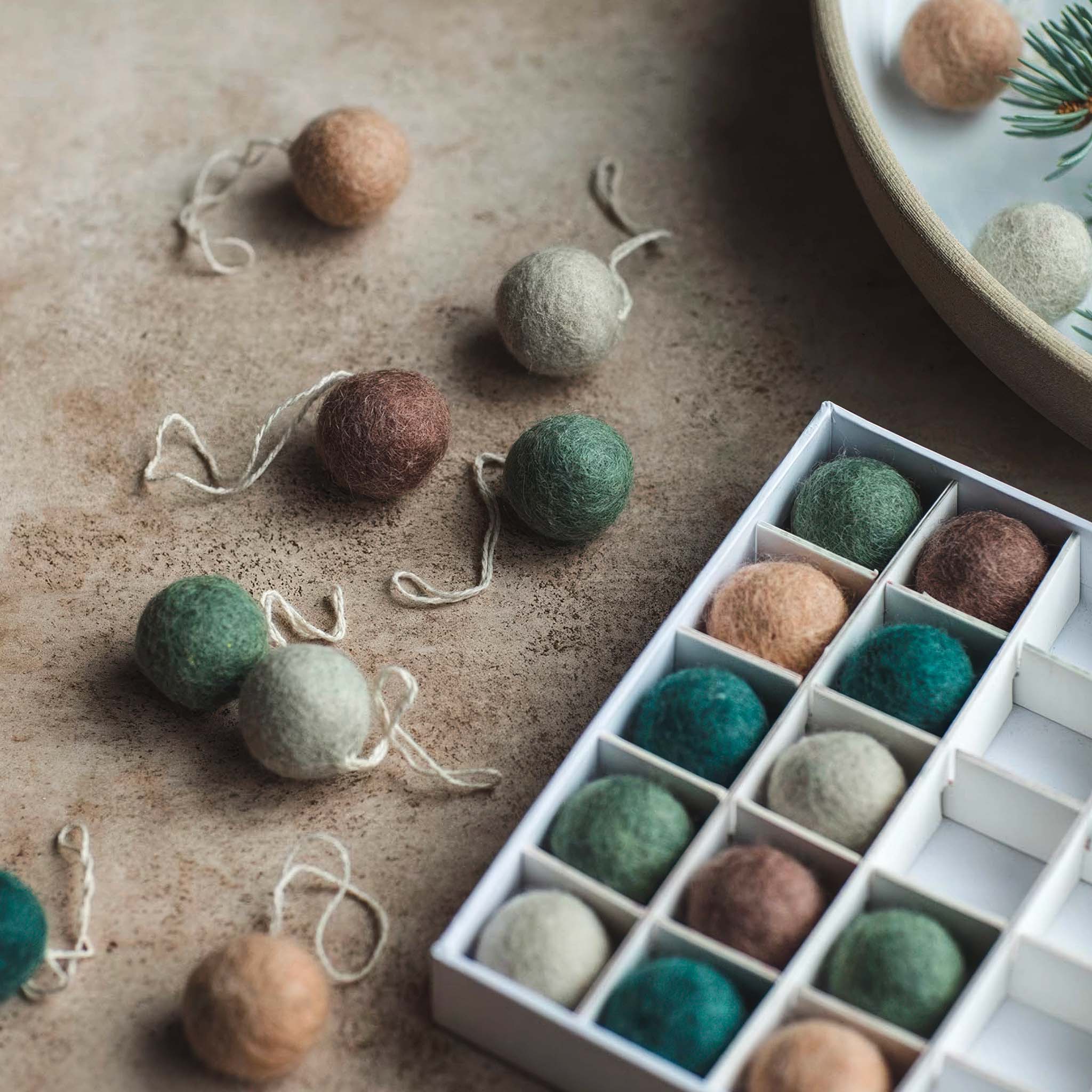 A set of felted wool baubles in earthy tones of green, teal, brown, blush, and cream are scattered on a textured surface beside an open box. The scene has a warm, natural feel with a few ornaments partially displayed in a ceramic dish with pine sprigs.