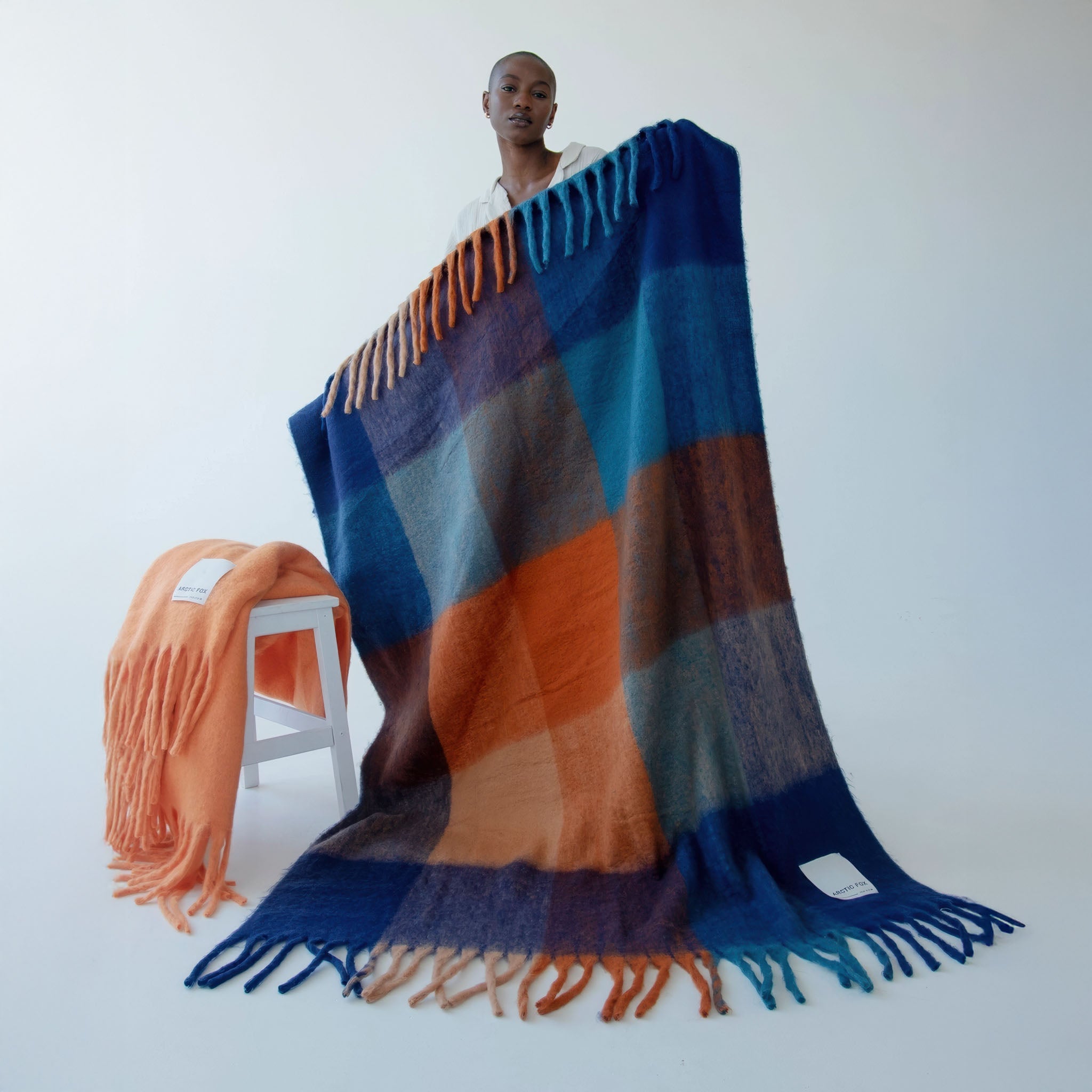 Model holding up a colorful plaid blanket with another blanket on a chair in the background.