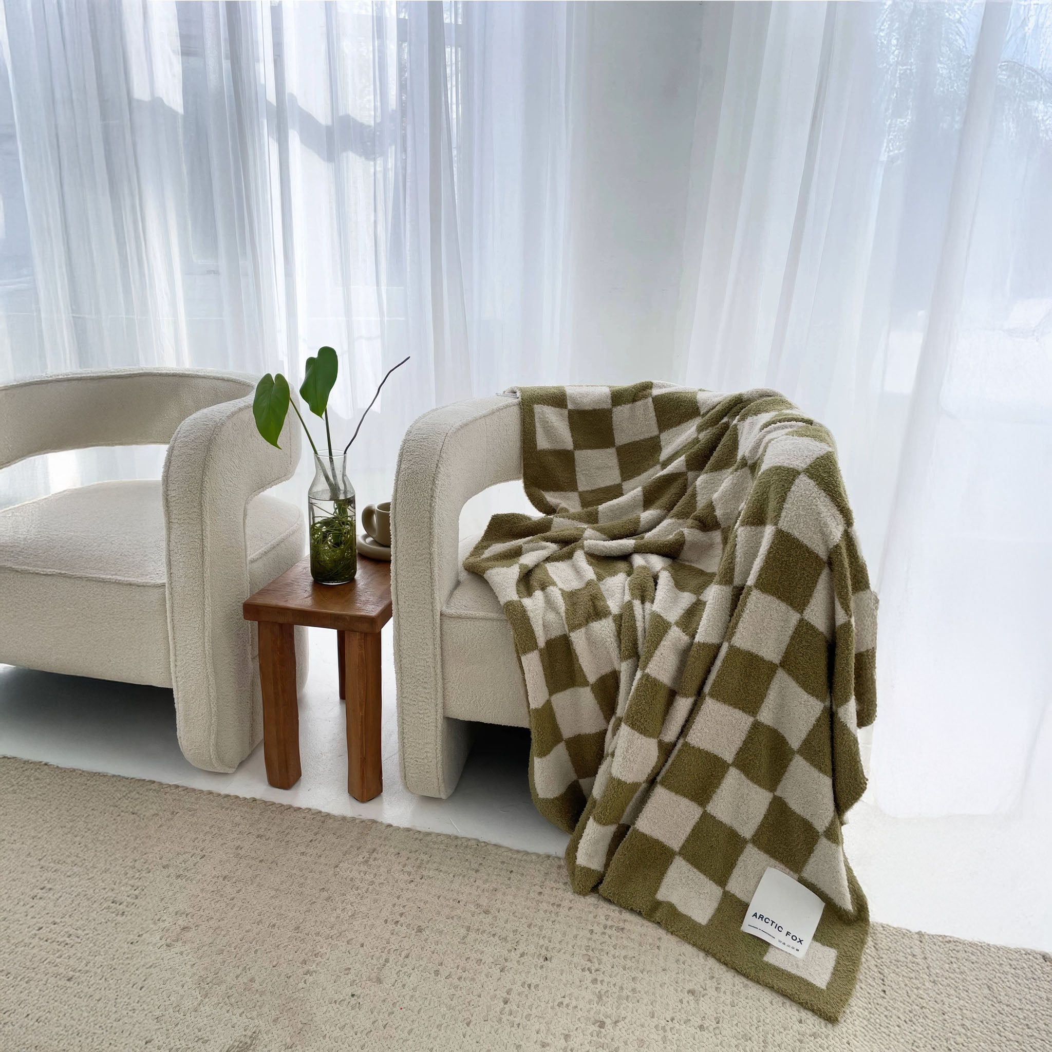 Moss green and white checkered throw blanket draped over a chair in a bright room with sheer curtains.