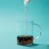 Full side view of the Multi-ccino mug with coffee while milk is being poured into the cup on a blue background