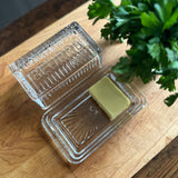 Block of butter in a glass container on a wooden surface with a plant in the background