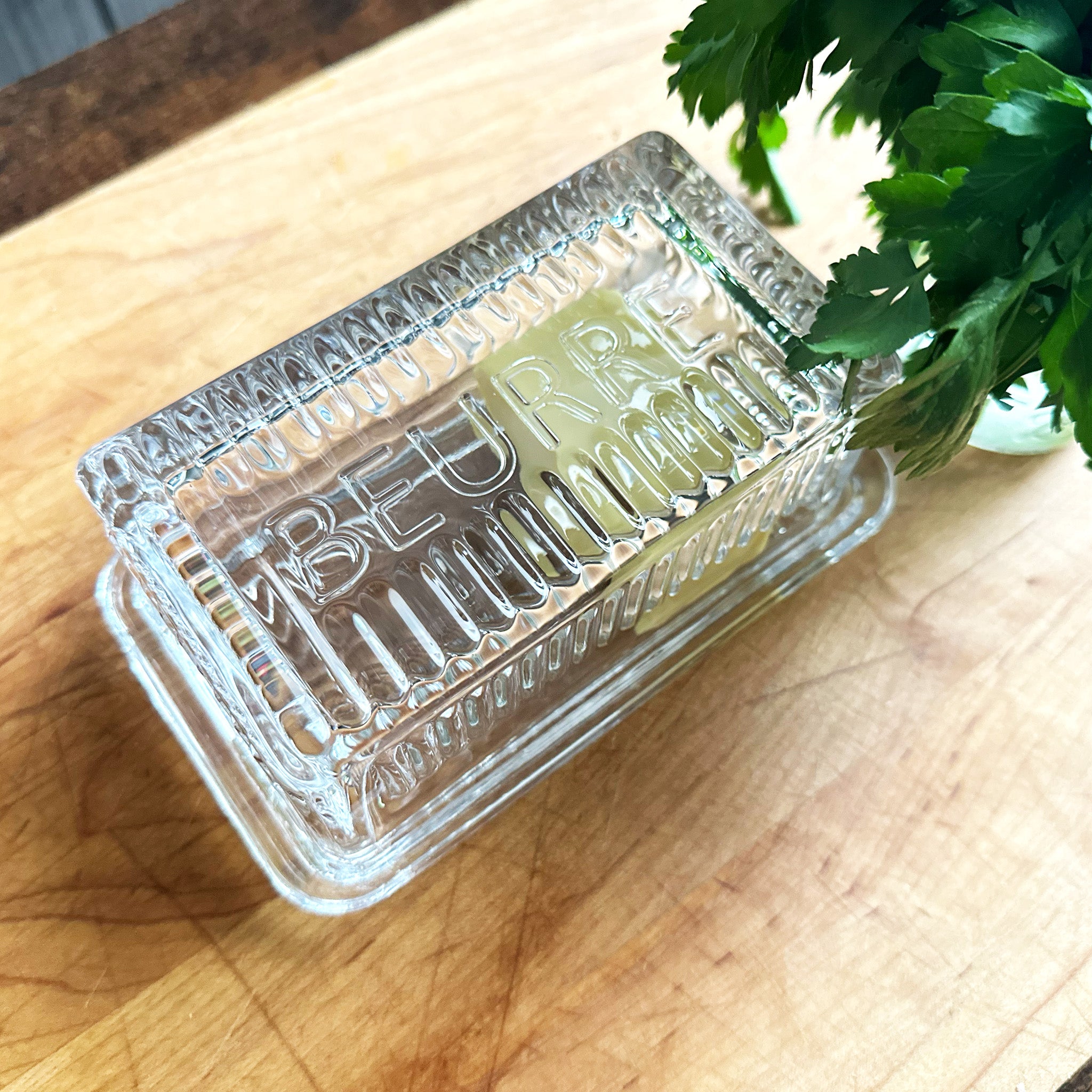 Clear glass beurre dish with embossed text on a wooden surface with a plant in the corner.