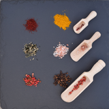Three wooden scoops filled with spices and seasonings on a dark slate background, surrounded by piles of turmeric, saffron, pink salt, sesame mix, peppercorns, and cloves.