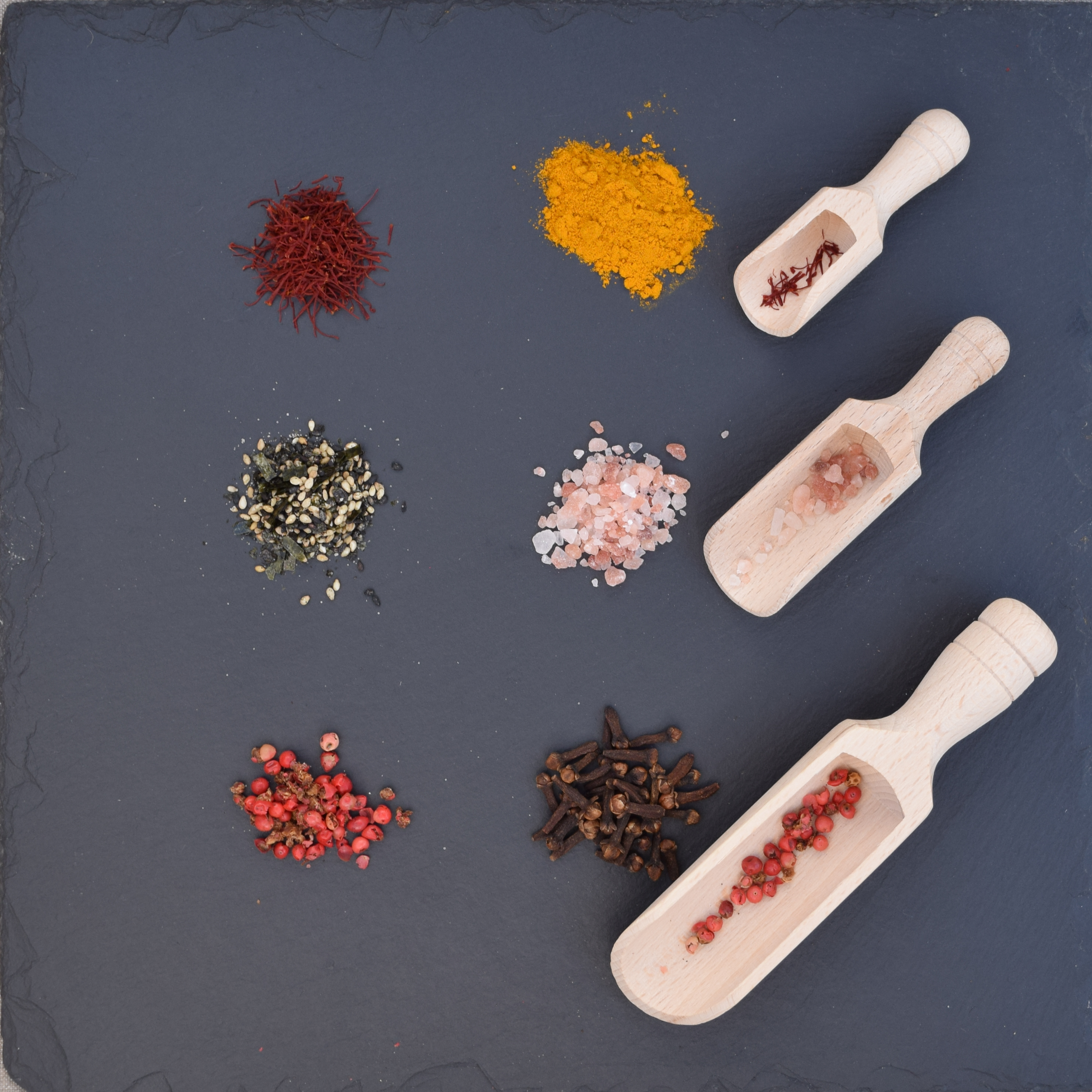 Three wooden scoops filled with spices and seasonings on a dark slate background, surrounded by piles of turmeric, saffron, pink salt, sesame mix, peppercorns, and cloves.