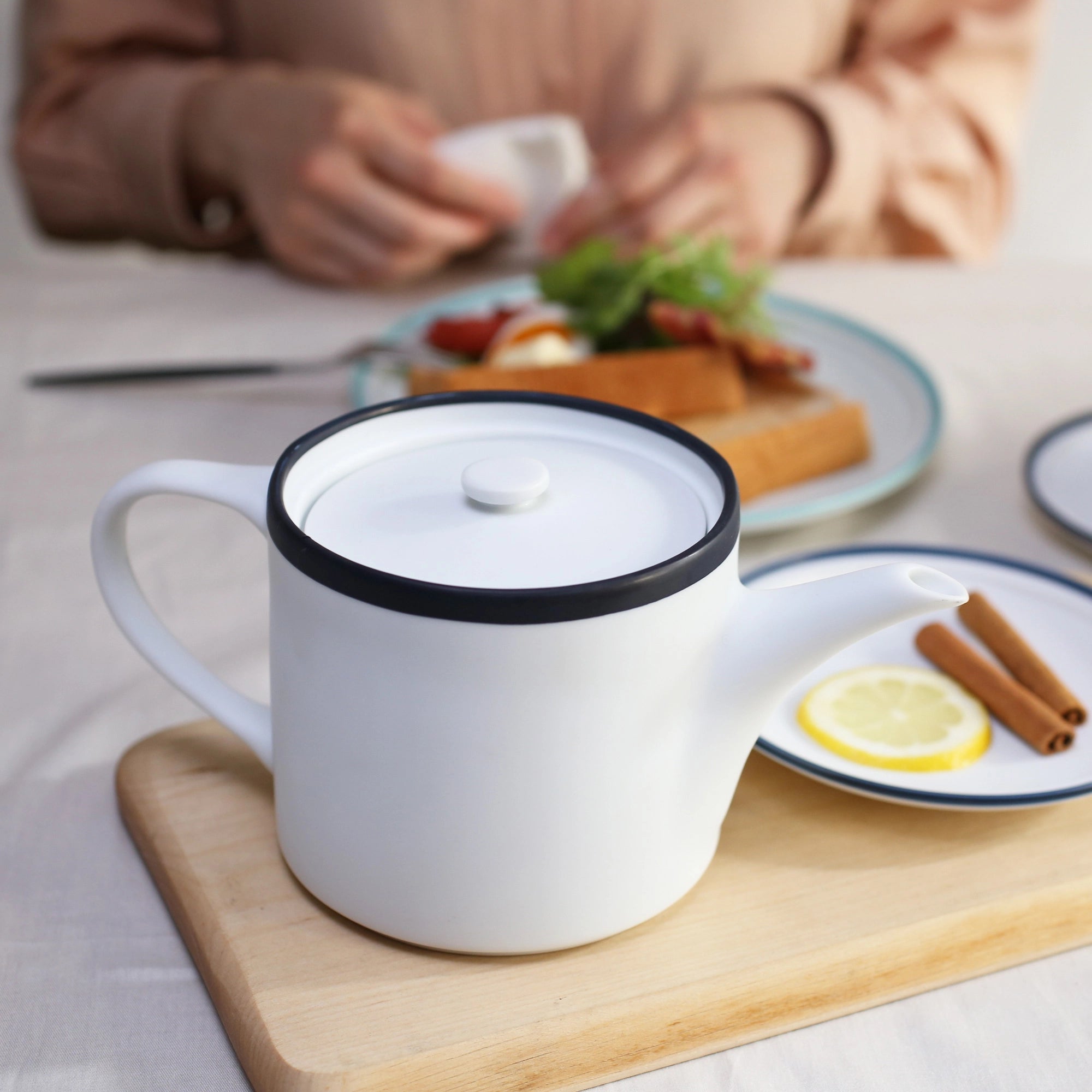 White teapot with black rim on a wooden cutting board, blurred background of a person eating.