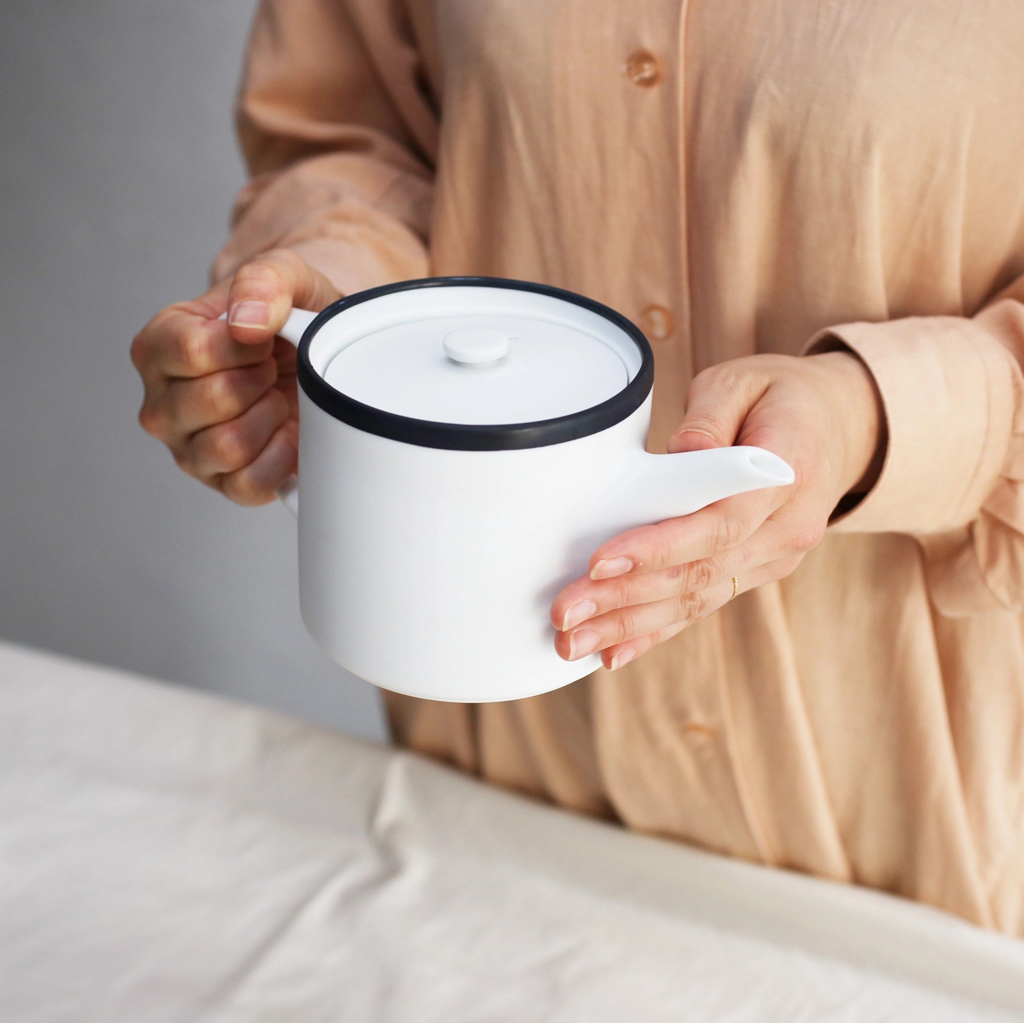 Person holding a white teapot with a black rim, wearing a beige shirt.