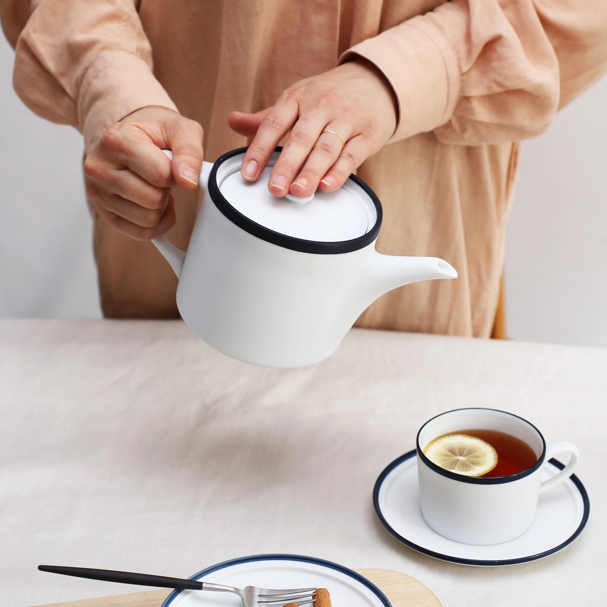 Person pouring tea from a white teapot into a cup with lemon on a light surface.