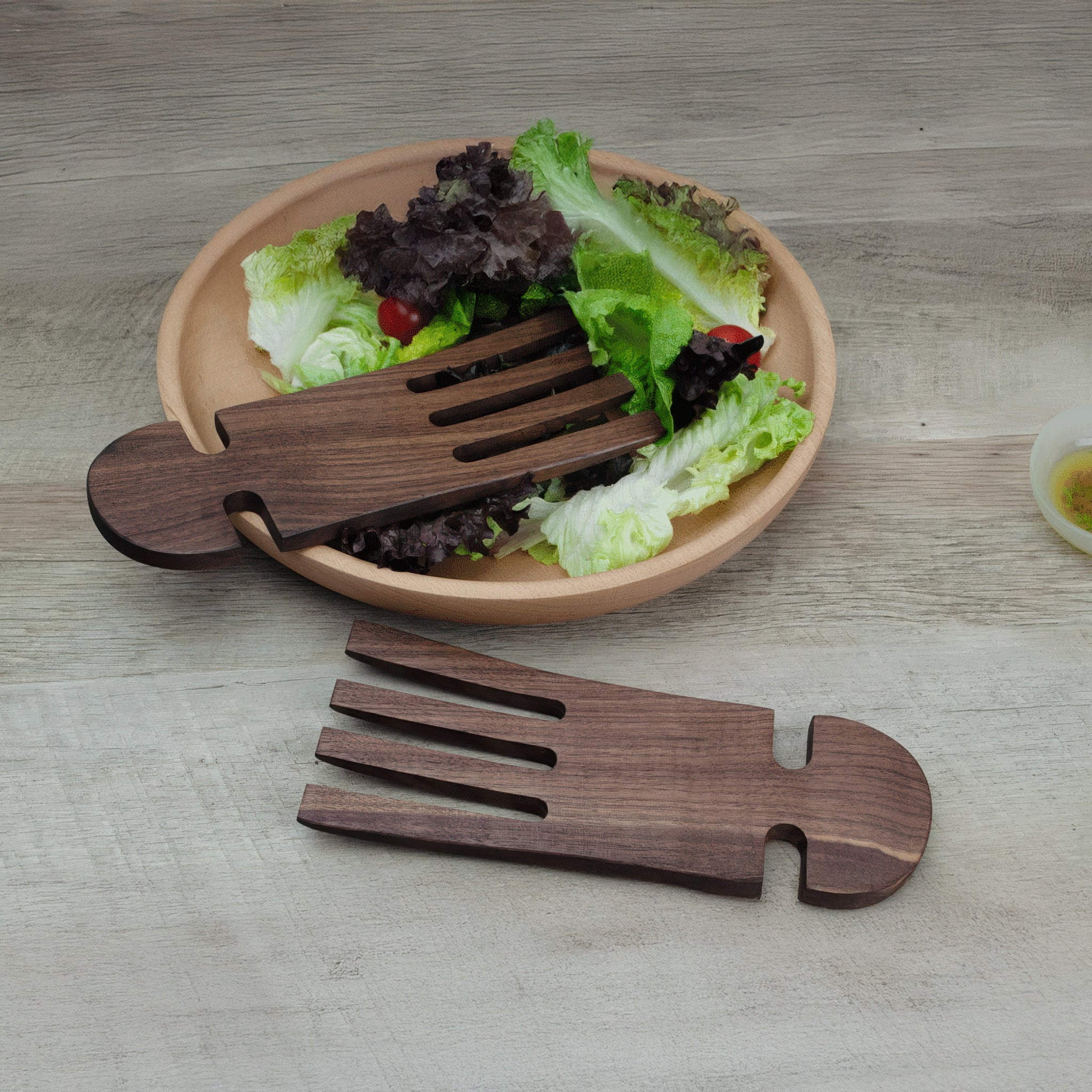 Dark walnut salad hands resting on a wooden table, one placed in a bowl of mixed greens and cherry tomatoes, the other beside it, with a small dish of dressing nearby.