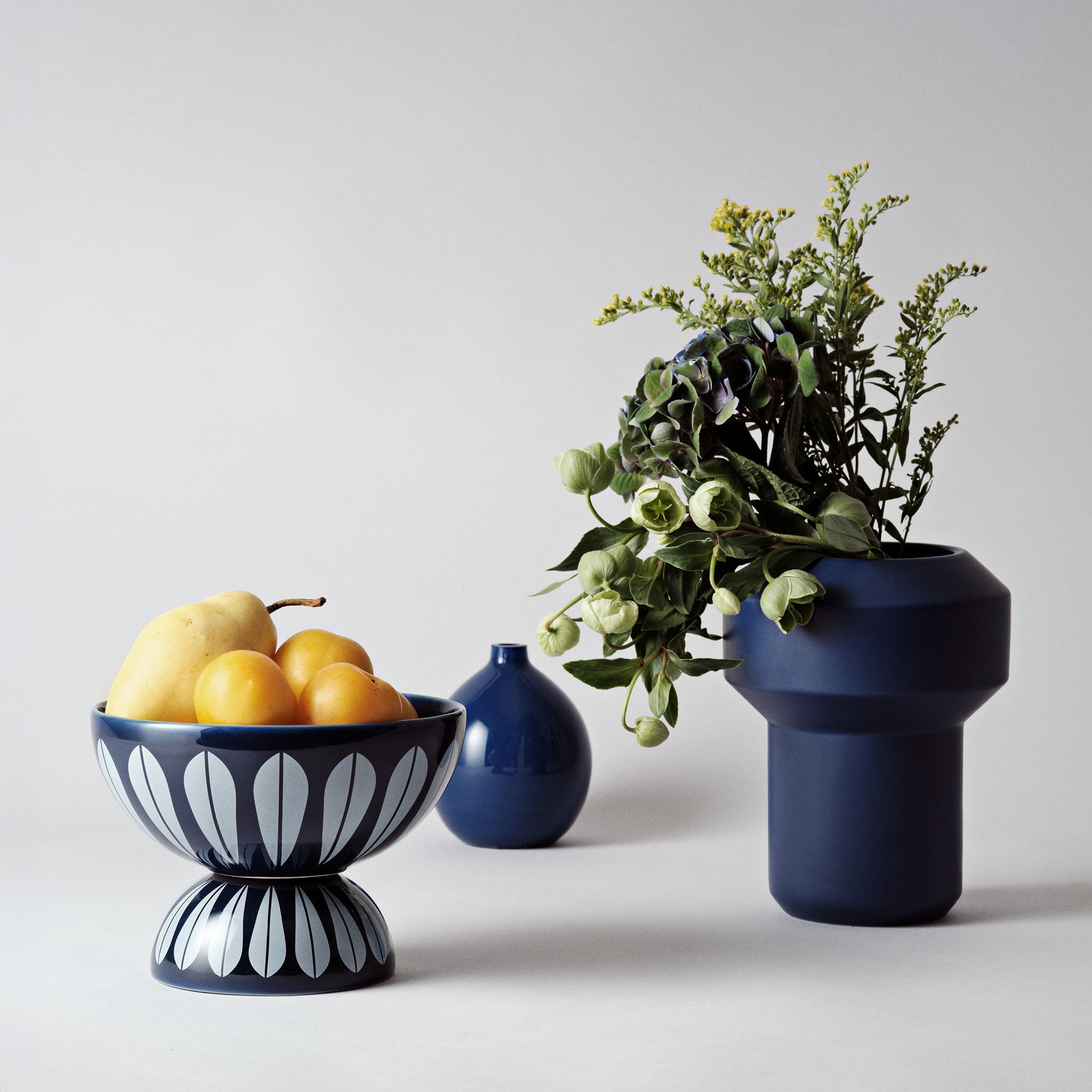 Blue Lucie Kaas Lotus Bowl stacked on inverted matching bowl, filled with yellow pears and plums, next to modern blue vases with green floral arrangement on a neutral backdrop.