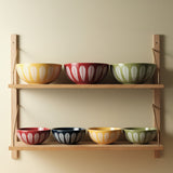 Assorted Lucie Kaas Lotus Bowls in red, yellow, green, and blue displayed on wooden shelves, showcasing a range of sizes and the iconic white lotus pattern.
