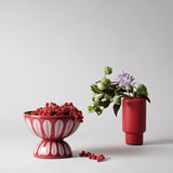Red Lucie Kaas Lotus Bowl stacked on matching inverted bowl, filled with red currants, beside a red vase holding green and purple flowers on a minimalist grey background.