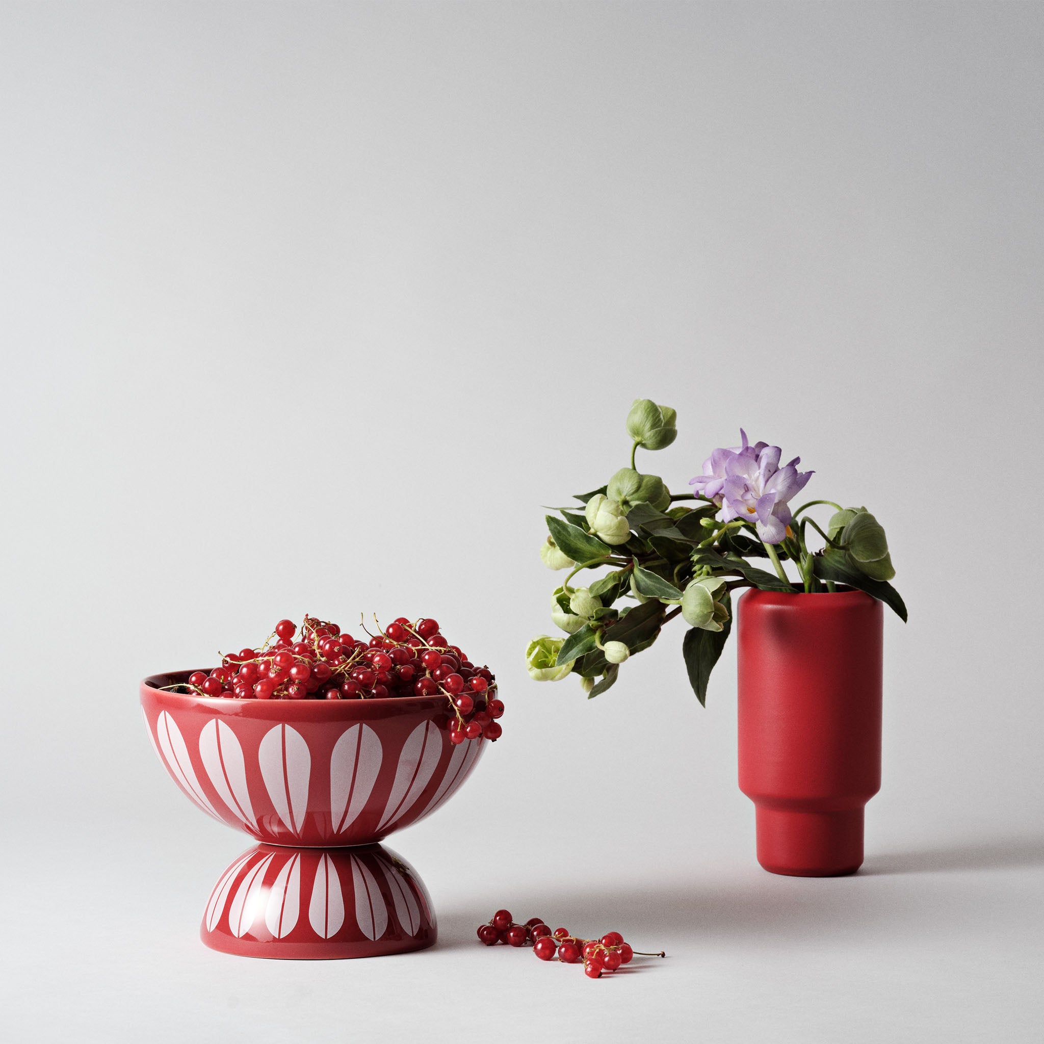 Red Lucie Kaas Lotus Bowl stacked on matching inverted bowl, filled with red currants, beside a red vase holding green and purple flowers on a minimalist grey background.