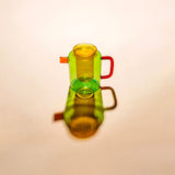 Colorful glass teapot with amber lid, yellow infuser, light green body, and red handle, shown against an amber background
