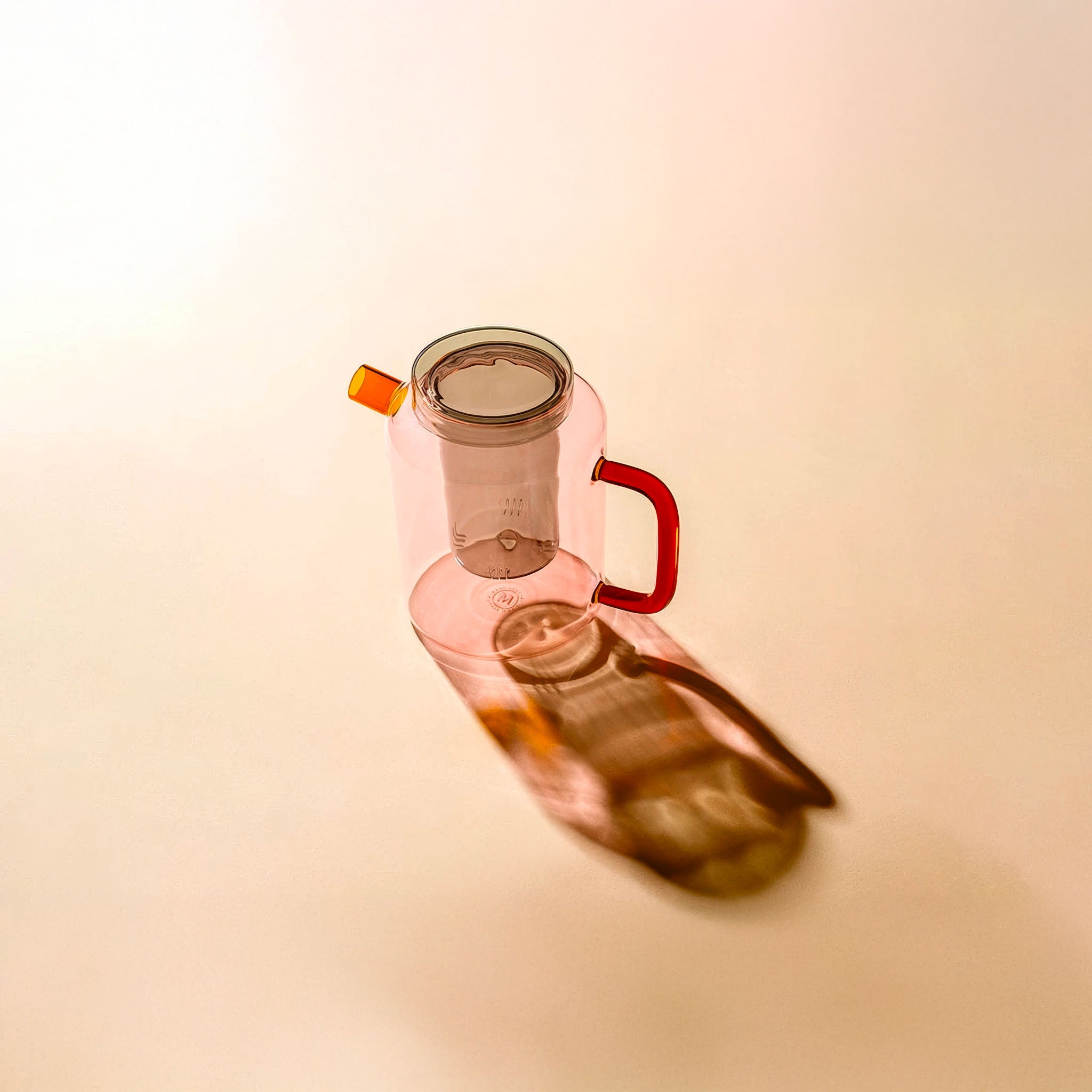 Pink glass teapot with amber spout, red handle, and clear gray lid and infuser, shown against a tan background.