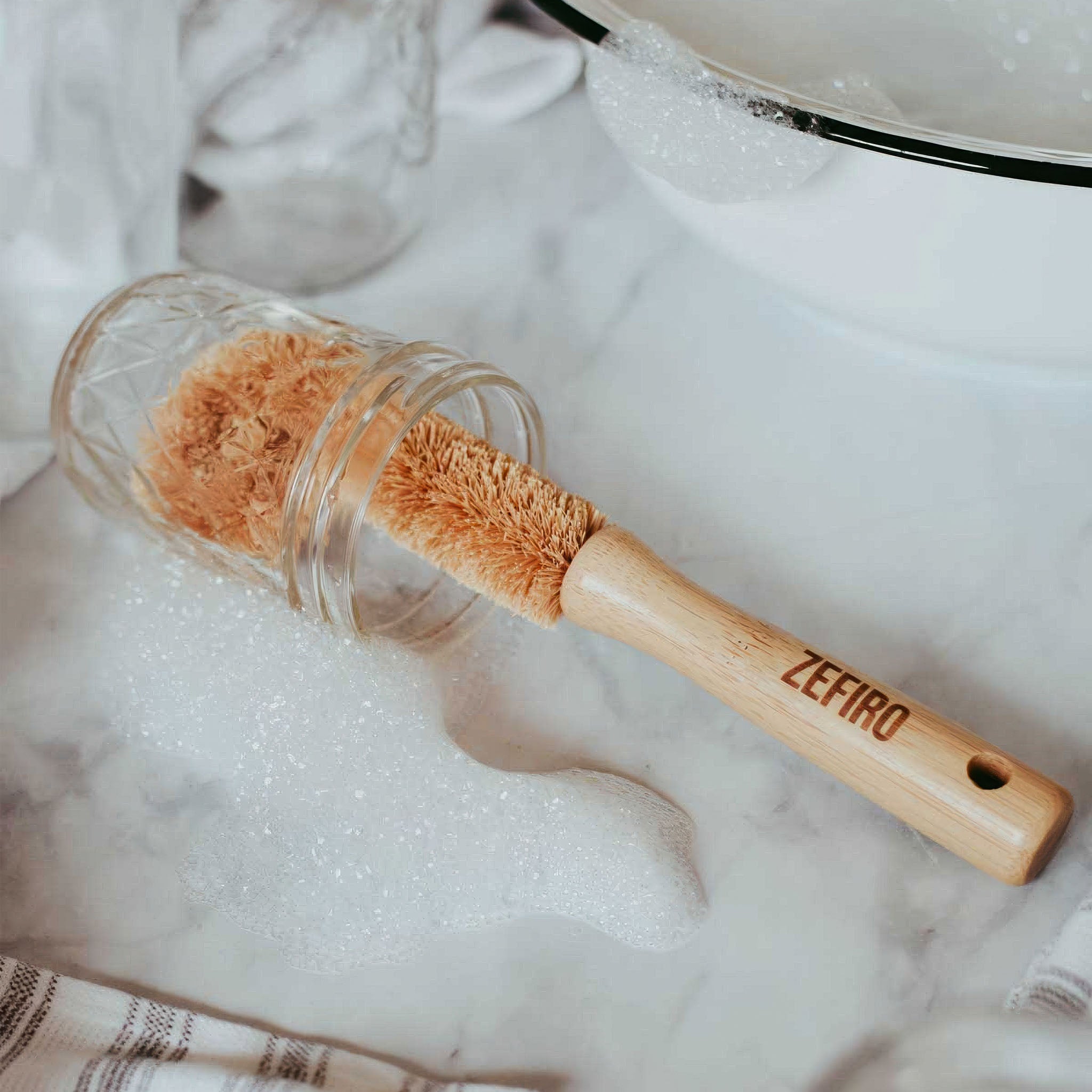 Glass bottle brush with wooden handle on a soapy surface, brand 'Zefiro' visible