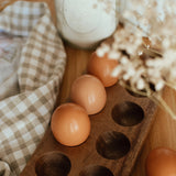 Brown eggs displayed in a wooden egg holder on a kitchen table with a gingham cloth and dried florals.