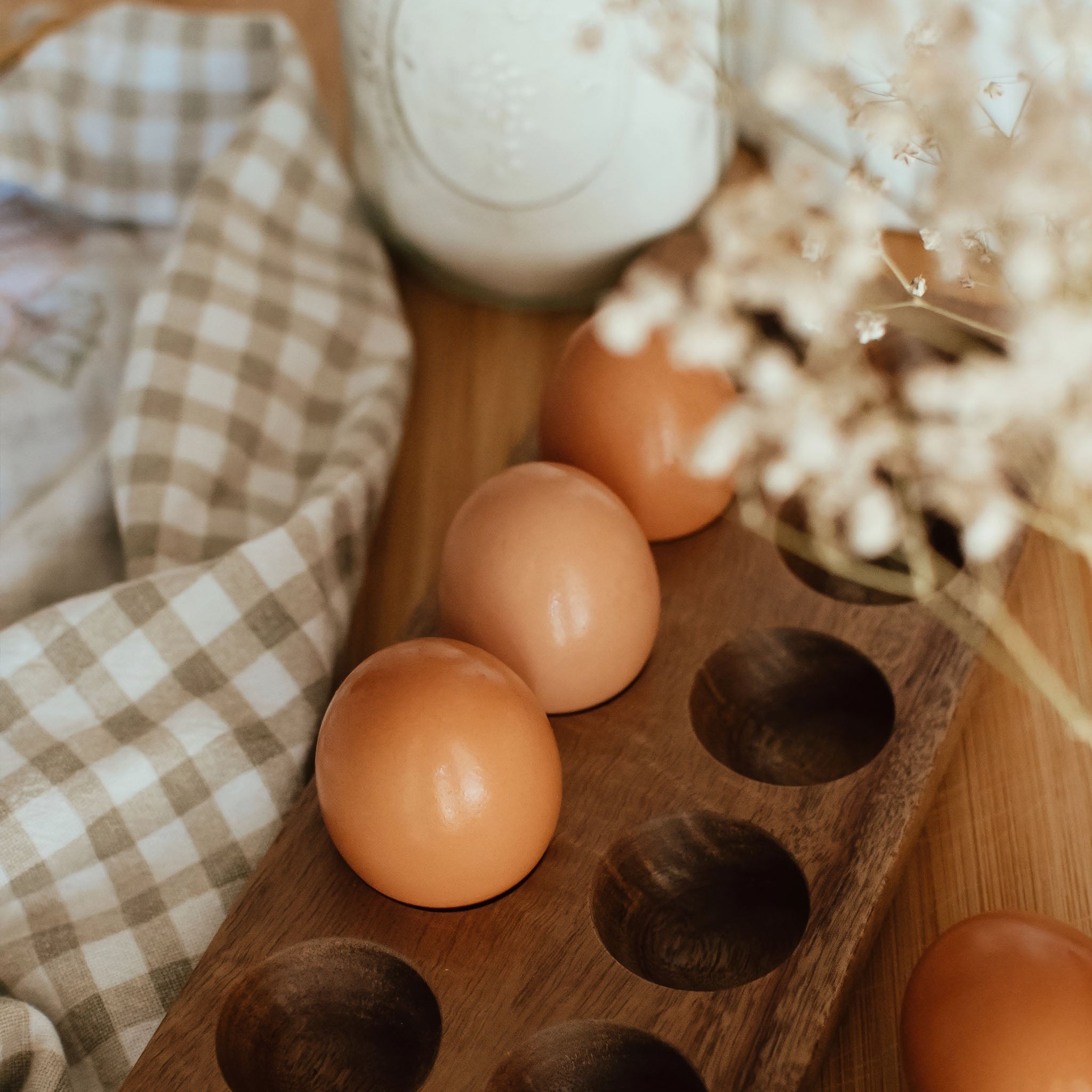 Brown eggs displayed in a wooden egg holder on a kitchen table with a gingham cloth and dried florals.
