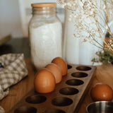 Wooden egg holder with fresh brown eggs on a kitchen counter beside a glass jar and soft neutral linens.
