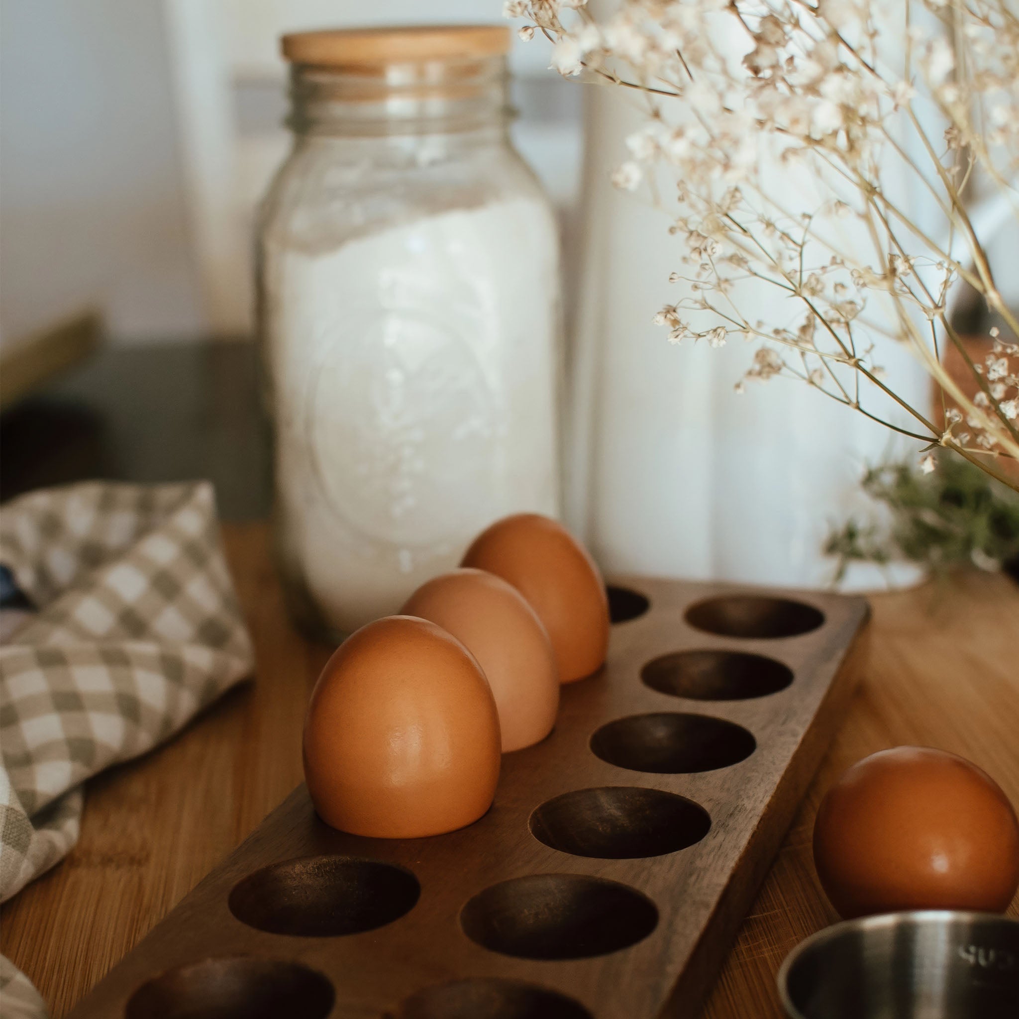 Wooden egg holder with fresh brown eggs on a kitchen counter beside a glass jar and soft neutral linens.