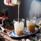 A hand pours a creamy drink from a glass bottle into two iced beverages on a rustic wooden table. The drinks are served in clear glasses placed on an oval ceramic tray with a blue floral pattern, surrounded by melting ice and soft natural light.
