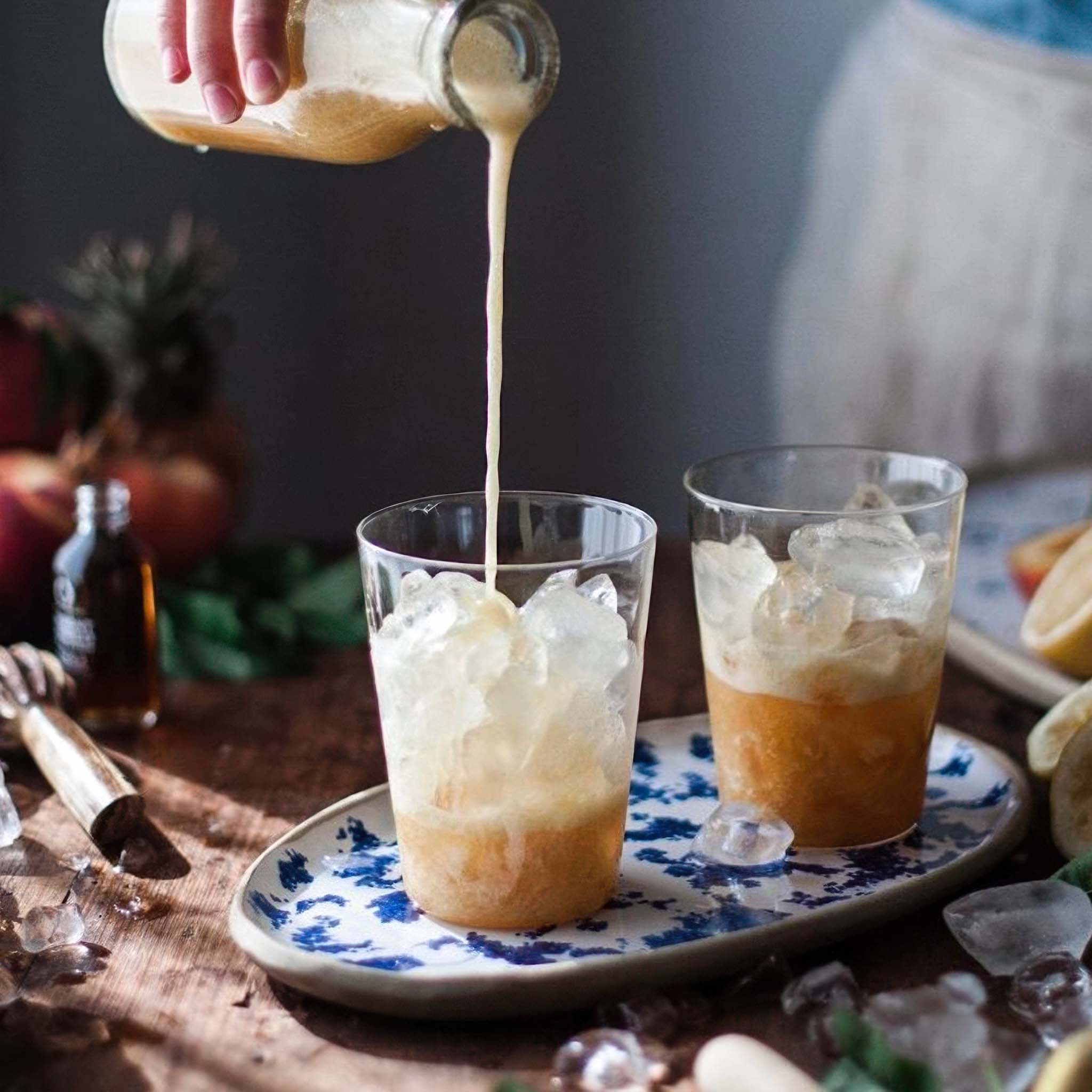 A hand pours a creamy drink from a glass bottle into two iced beverages on a rustic wooden table. The drinks are served in clear glasses placed on an oval ceramic tray with a blue floral pattern, surrounded by melting ice and soft natural light.