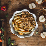 A white ceramic pie dish full of apple crisp on a wooden table with sliced apples around