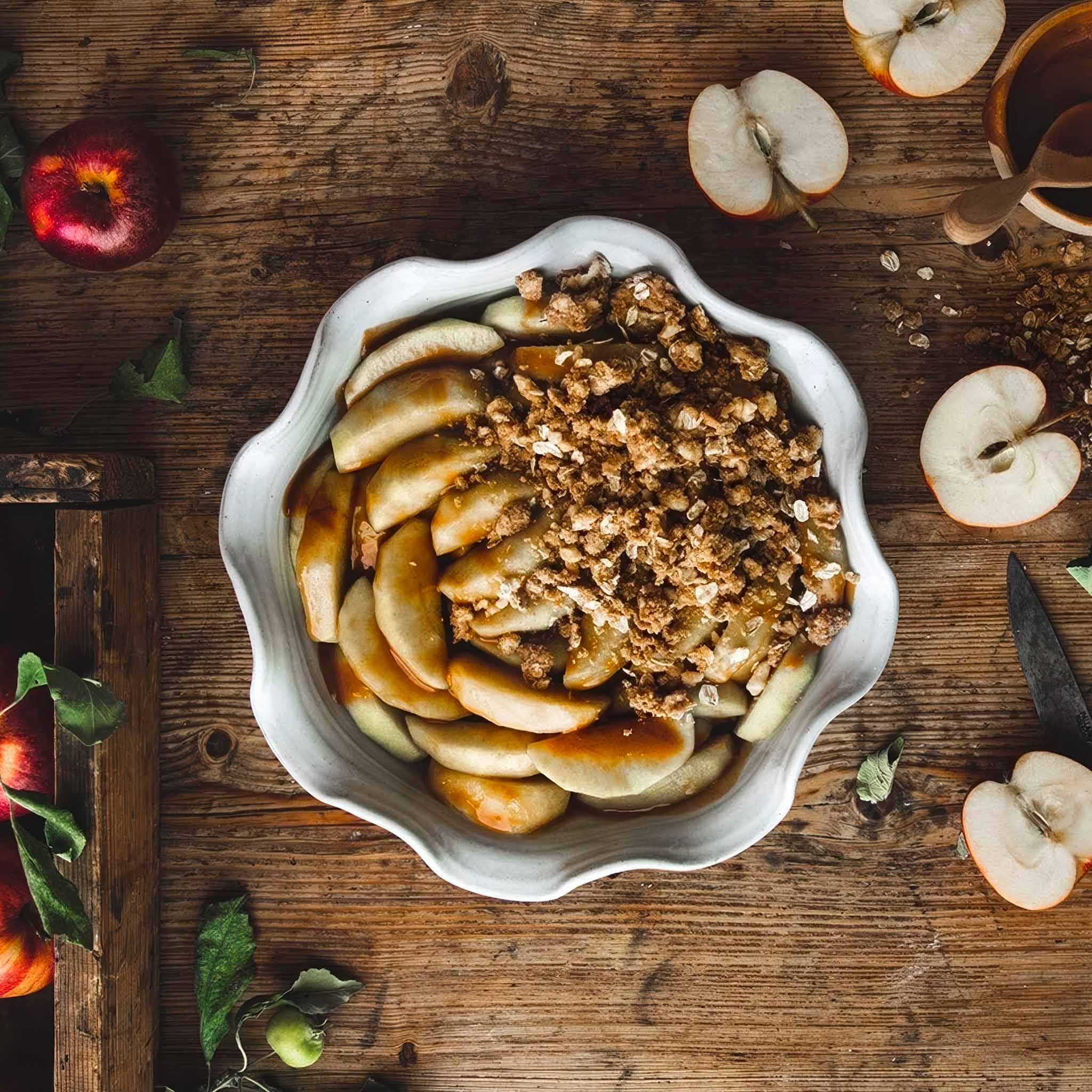 A white ceramic pie dish full of apple crisp on a wooden table with sliced apples around