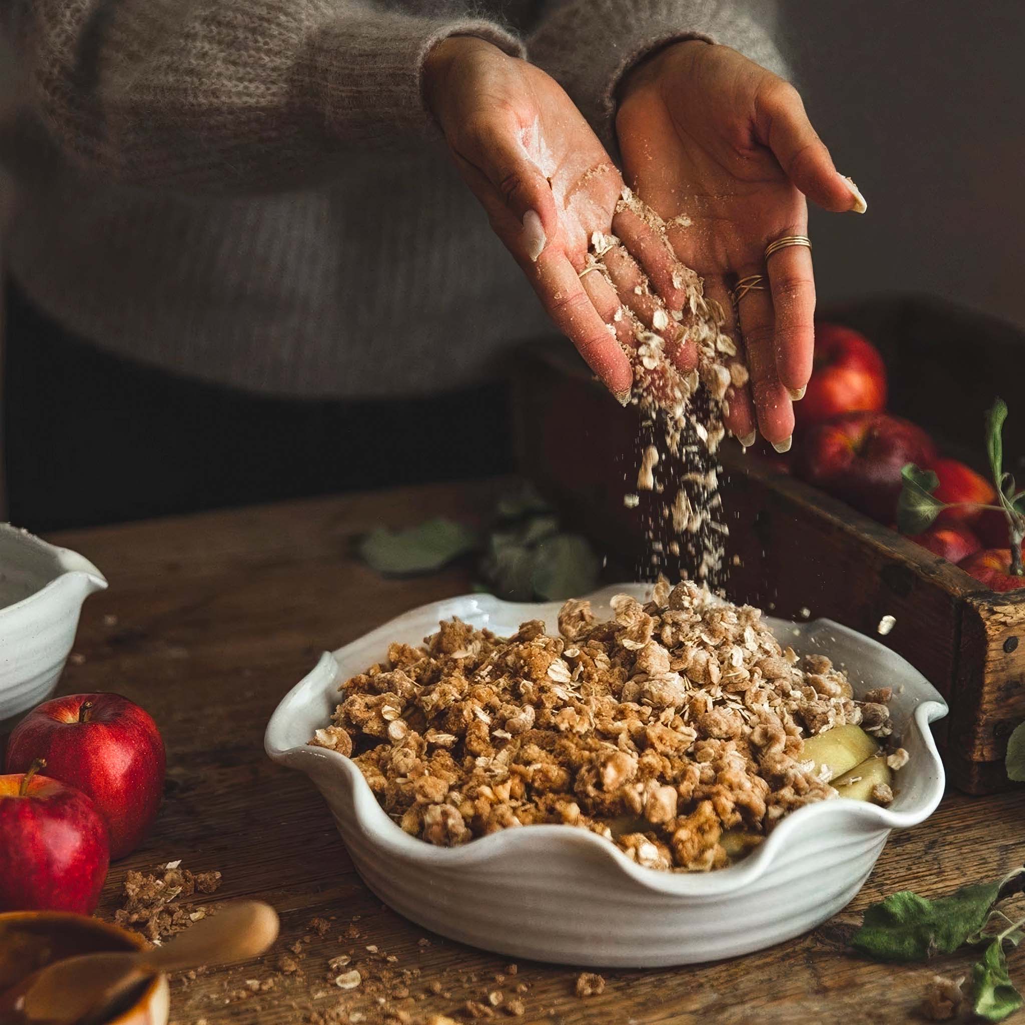 Person pouring crumble topping onto a white ceramic pie dish with apples on a wooden table
